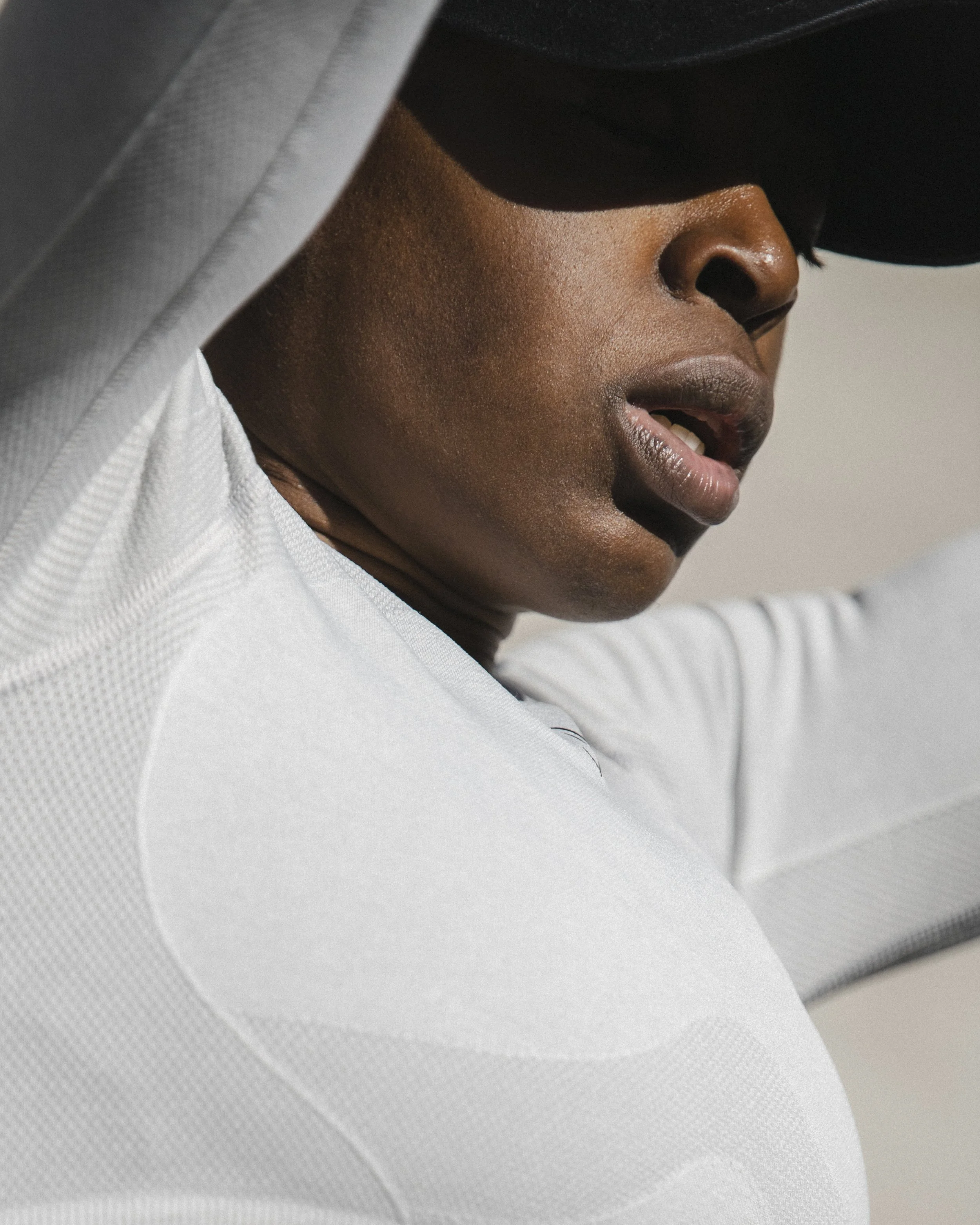 Close-up of a person wearing a white sports shirt, their face partially covered by a black hat or visor, with visible nose, lips, and cheek.