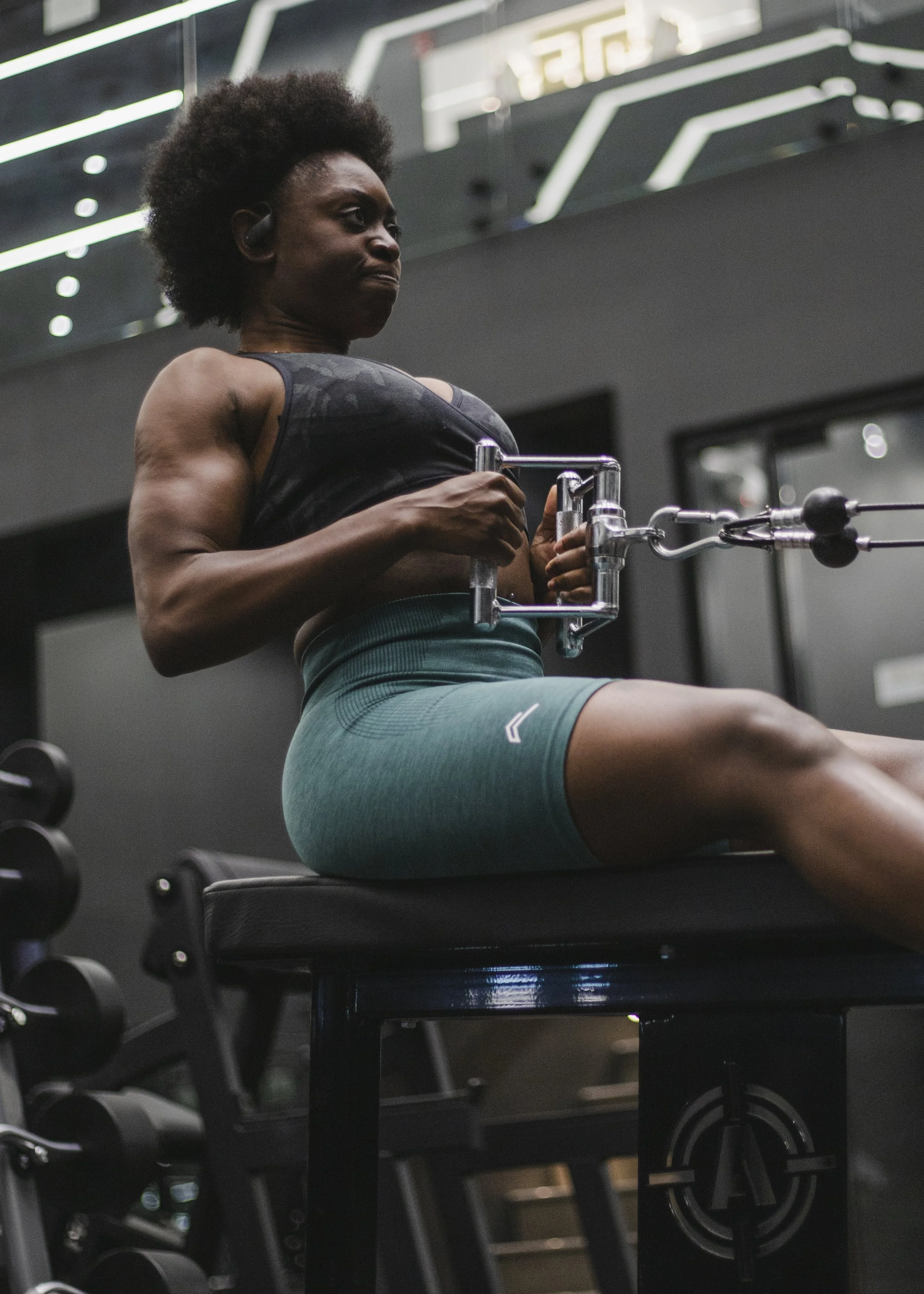 A woman with an afro hairstyle performing a seated tricep extension exercise at the gym, wearing headphones, a black tank top, and teal workout shorts, with gym equipment and mirrors in the background.