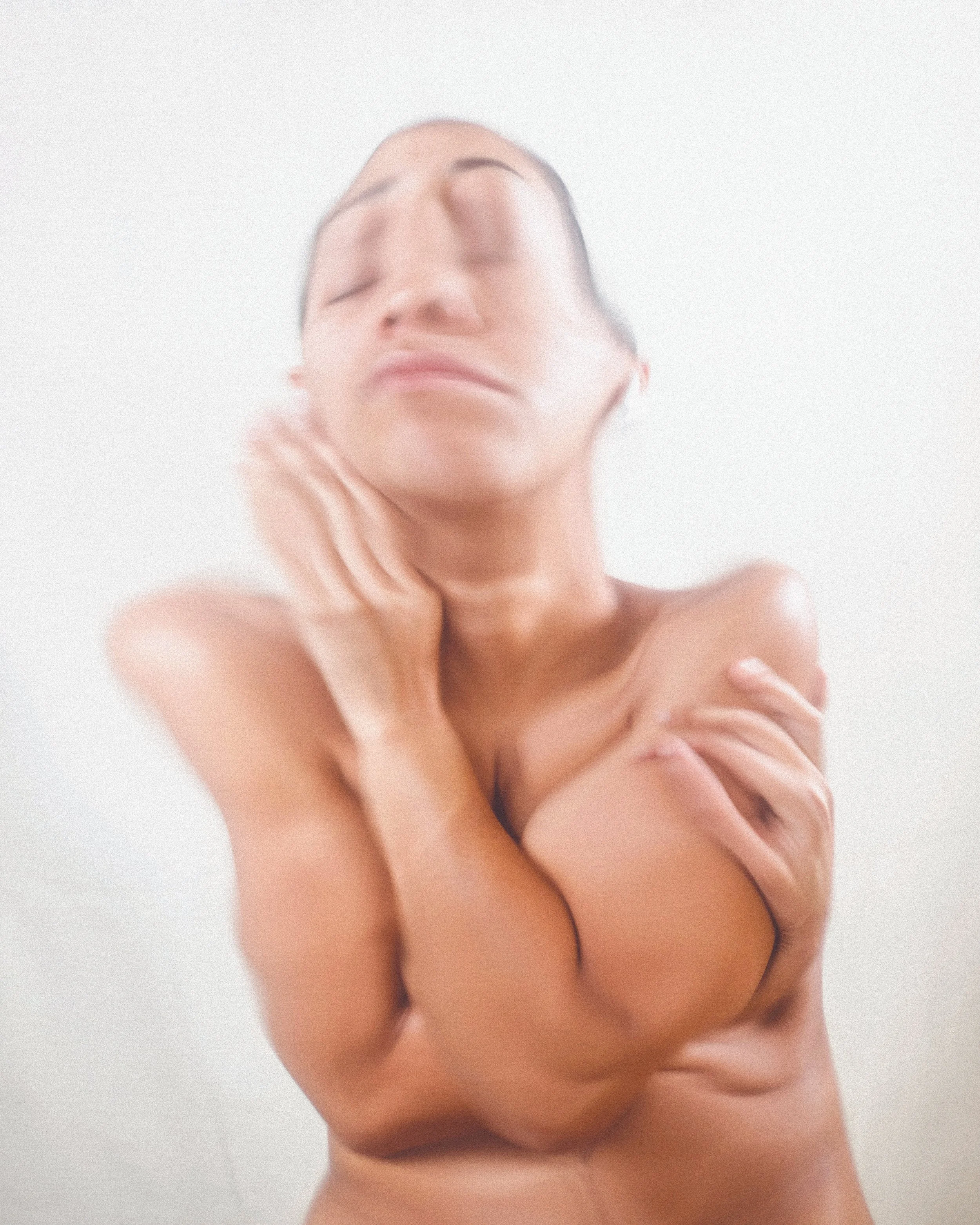 A nude woman with closed eyes, touching her neck and shoulder against a plain white background.