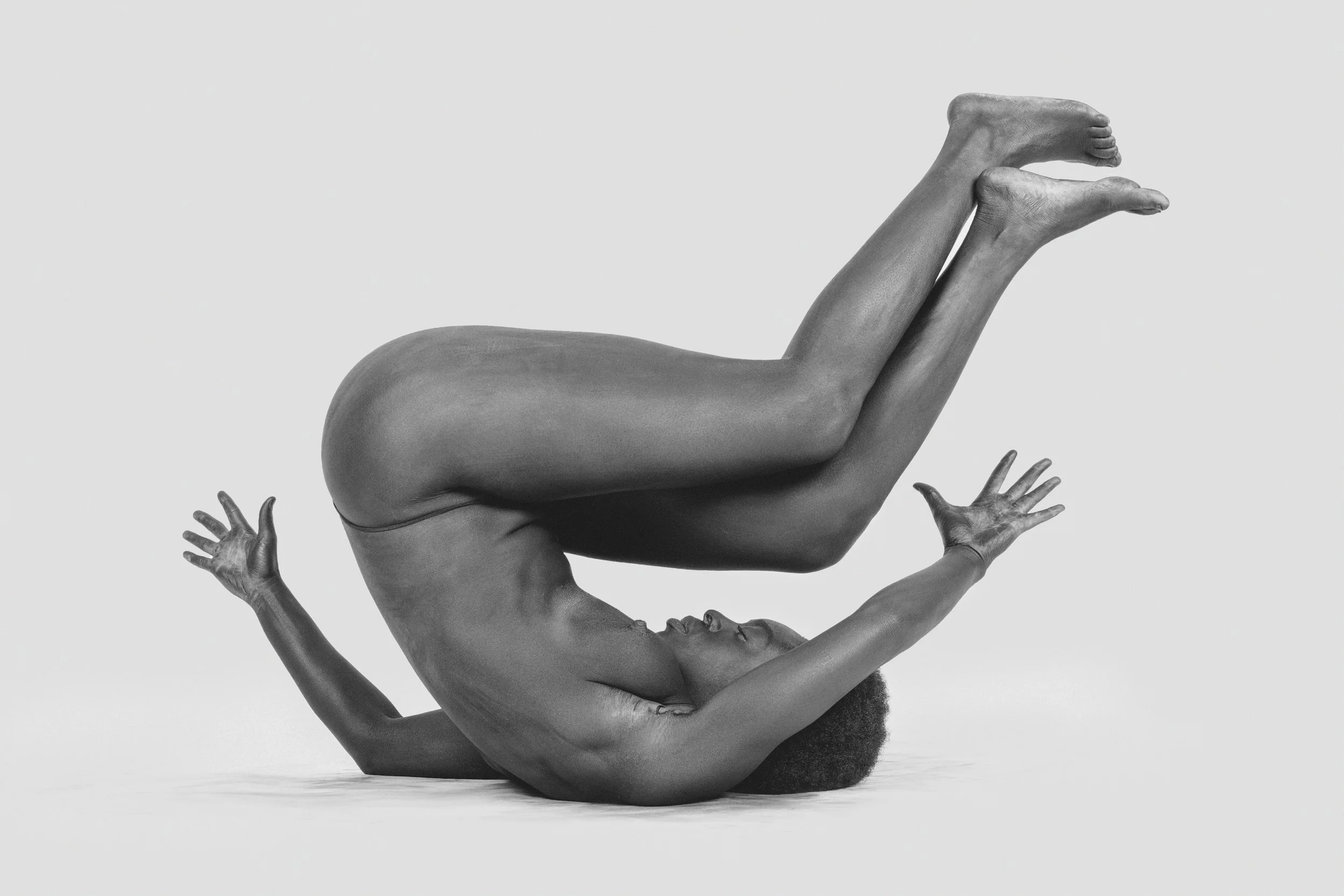 A black and white photograph of a human figure in an unusual pose, with their upper body reversed and arms extended upward, supporting their body. The head and face are upside down, and the hands are reaching out with fingers spread open.