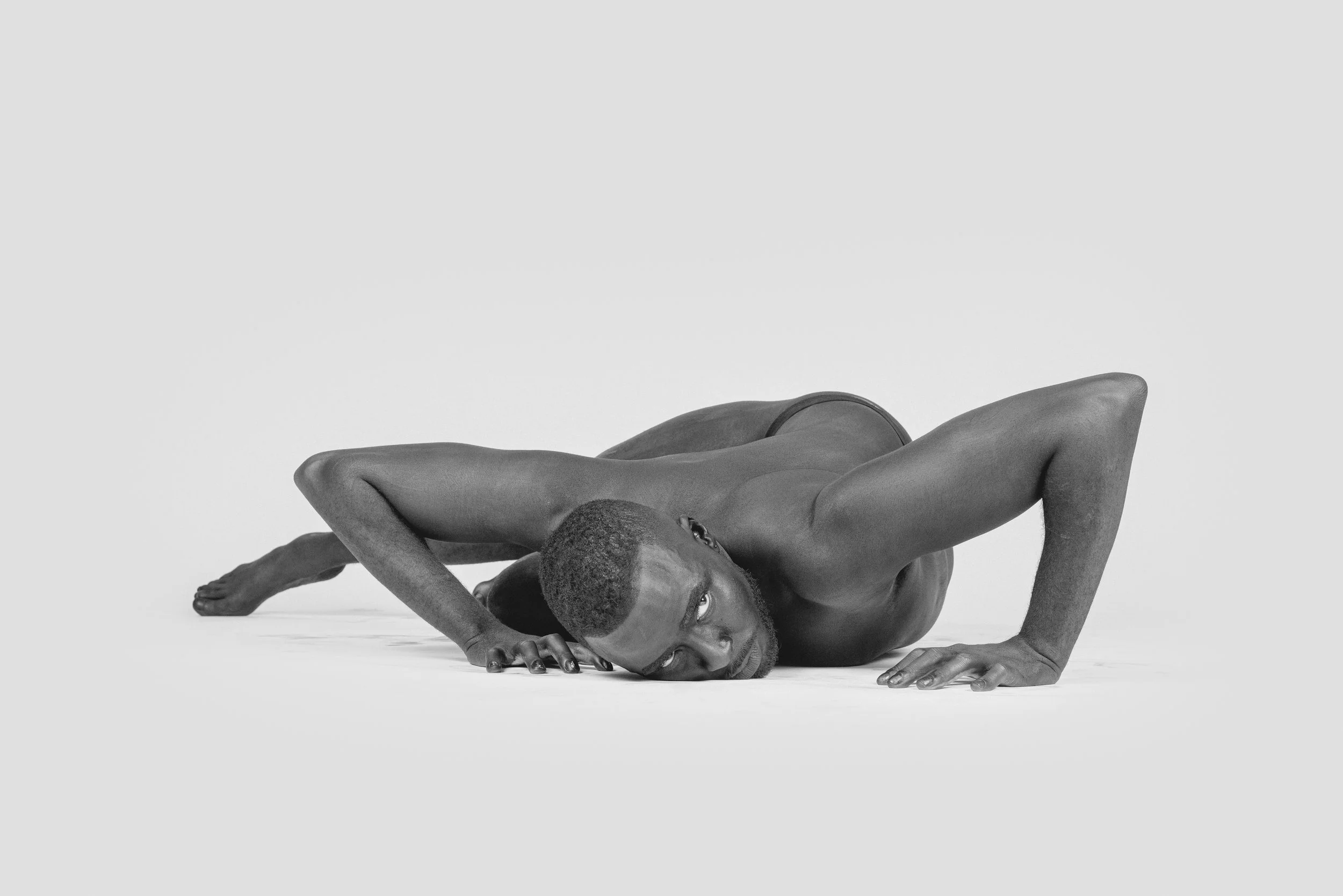 A black and white photo of a person with short hair, in a crouched position, supporting their head with one hand and looking into the camera with an intense gaze.