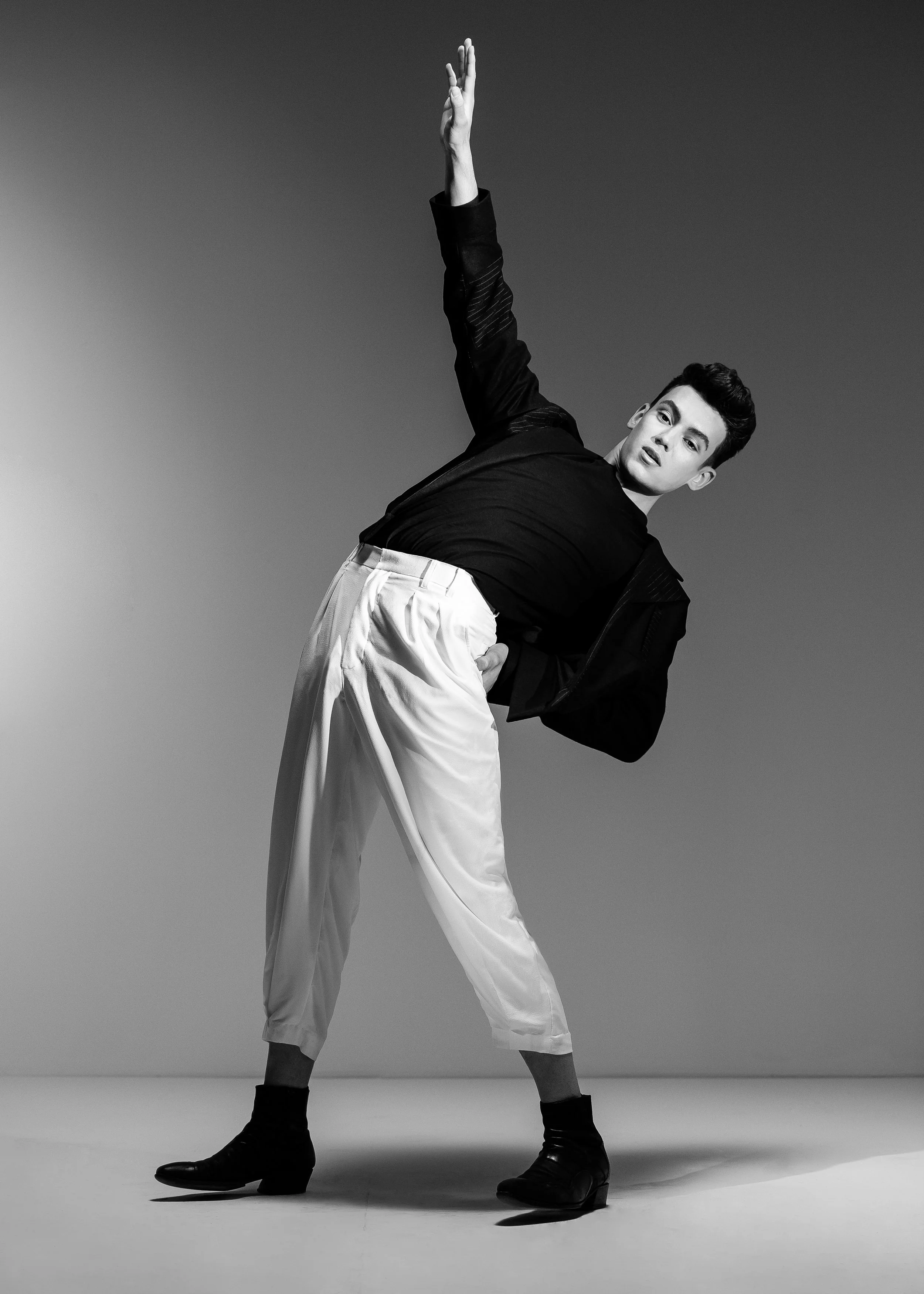 Black and white photo of a young man posing with one arm raised, wearing a black jacket, black shirt, light-colored pants, black socks, and black boots.