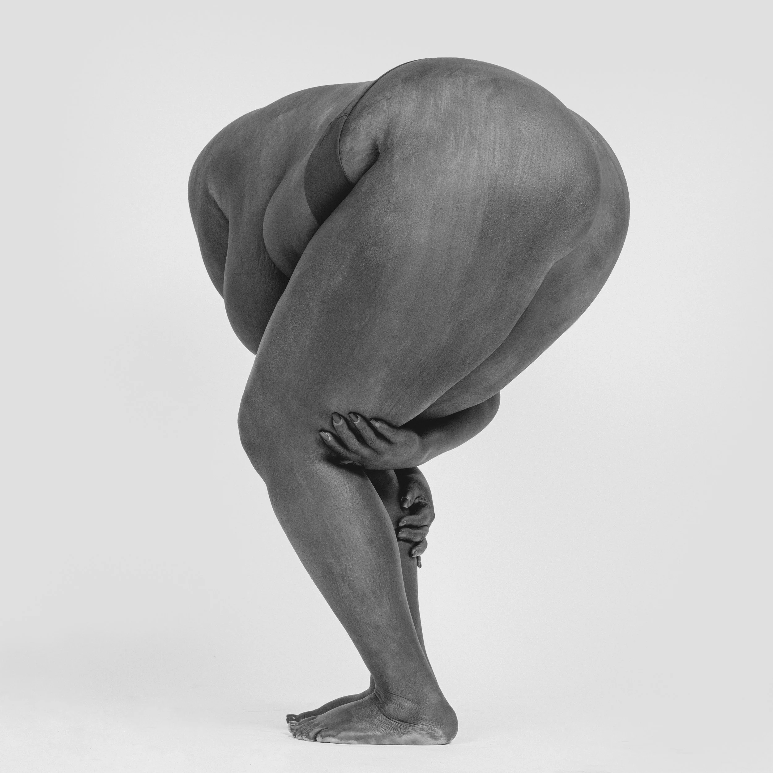 Black and white photograph of a human figure in a yoga pose, with arms crossed and hands resting on ankles, viewed from the back, against a plain light background.