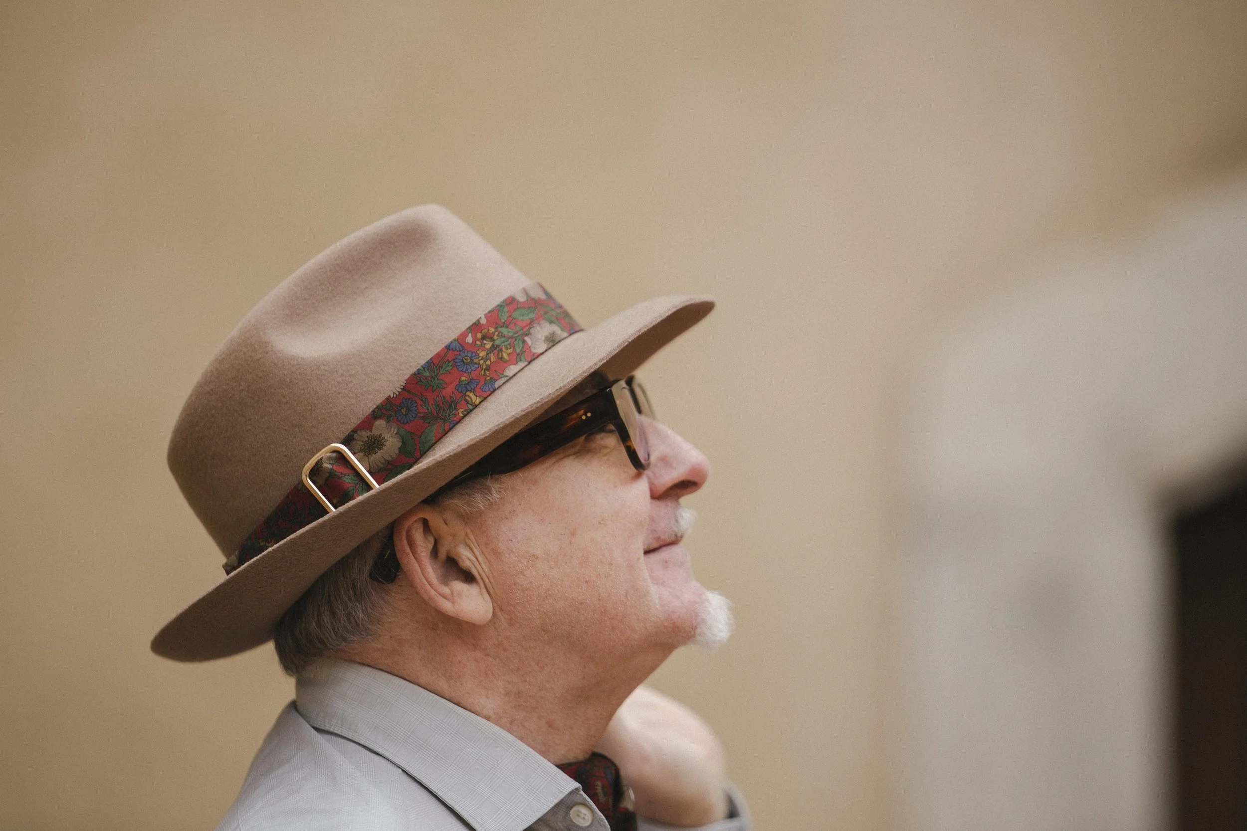 A side profile of an elderly man wearing glasses, a light gray dress shirt, a beige hat with a floral band, and a small portion of a necktie visible.
