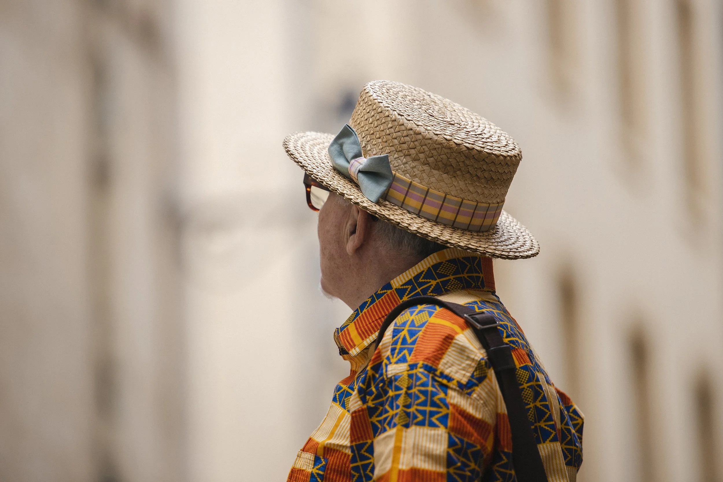 A person wearing a woven straw hat with a pastel-colored ribbon and bow, a colorful patterned shirt, and a backpack, walking in an indoor space with blurred beige walls.