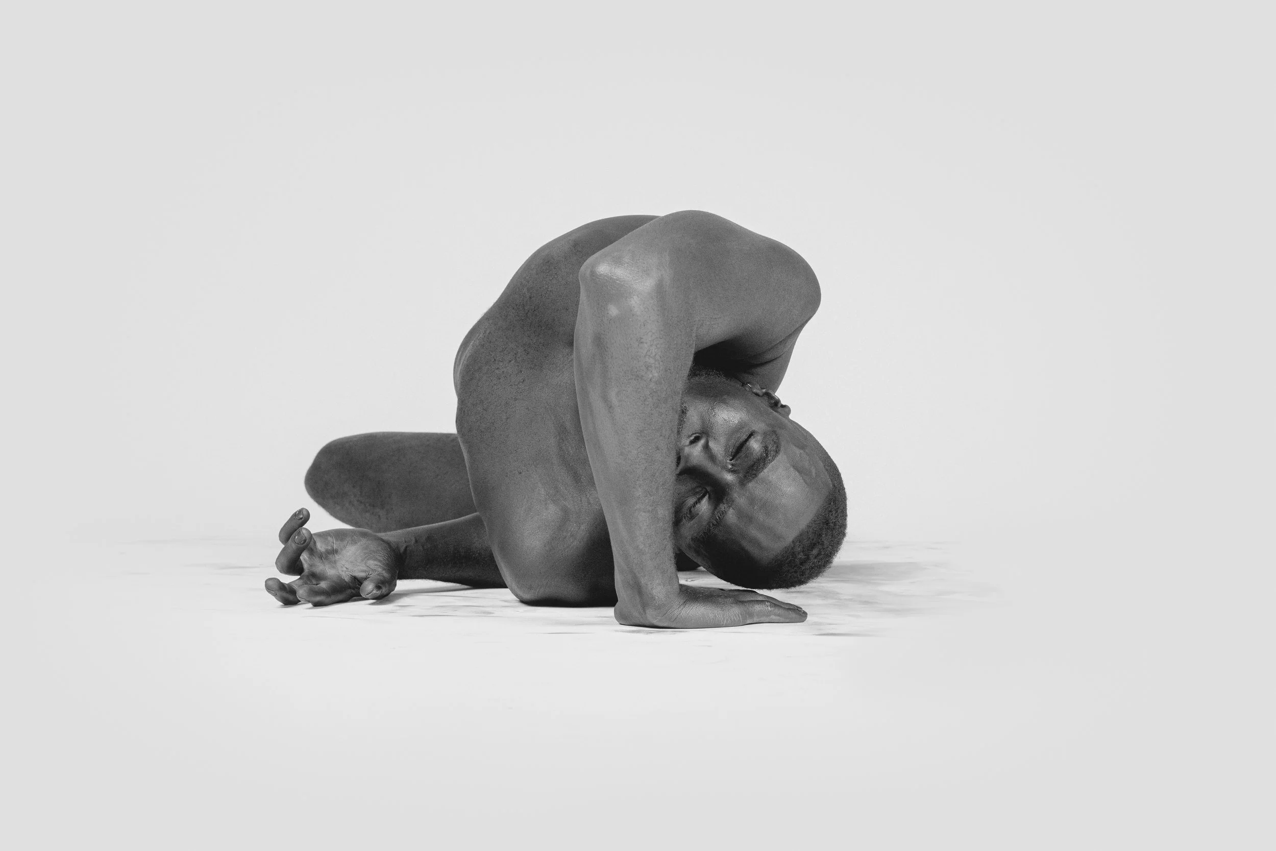 A man with a shaved head and a beard is practicing yoga, sitting on the floor with one leg extended and bent at the knee, and the other leg folded under him. He is leaning forward, supporting his body with one arm on the ground, and his other arm is 