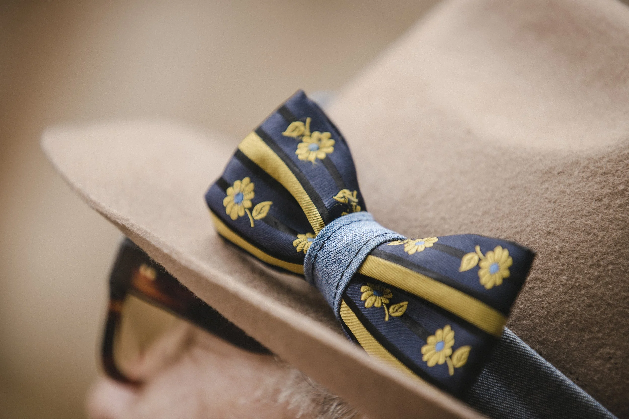 Close-up of a pair of blue denim jeans with a flower-printed navy blue and yellow fabric bow on the ankle, resting on a beige upholstered surface.