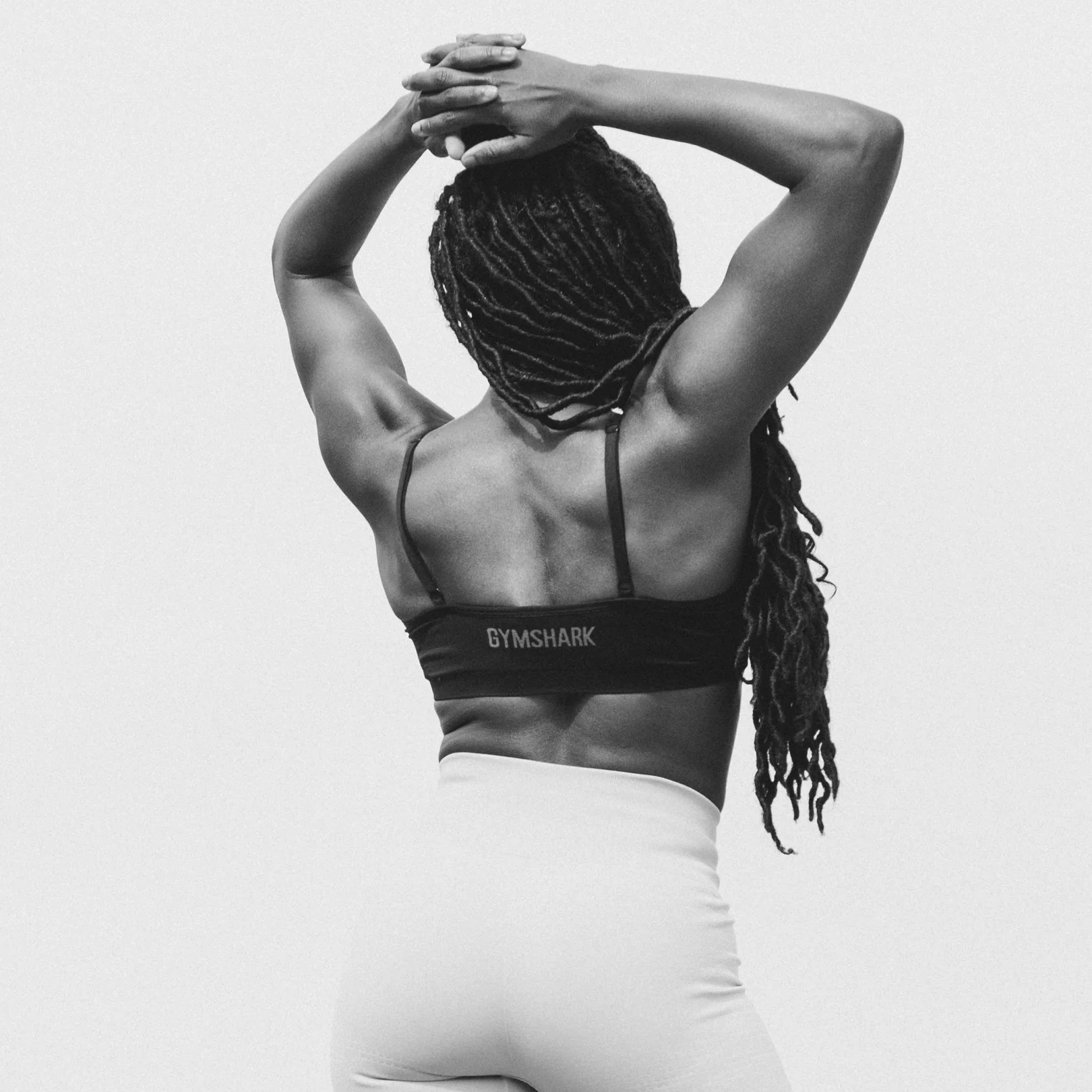 Black and white photo of a woman with twisted dreadlocks stretching over her shoulder, wearing a Gymshark sports bra and high-waisted leggings, with arms raised and hands clasped behind her head.
