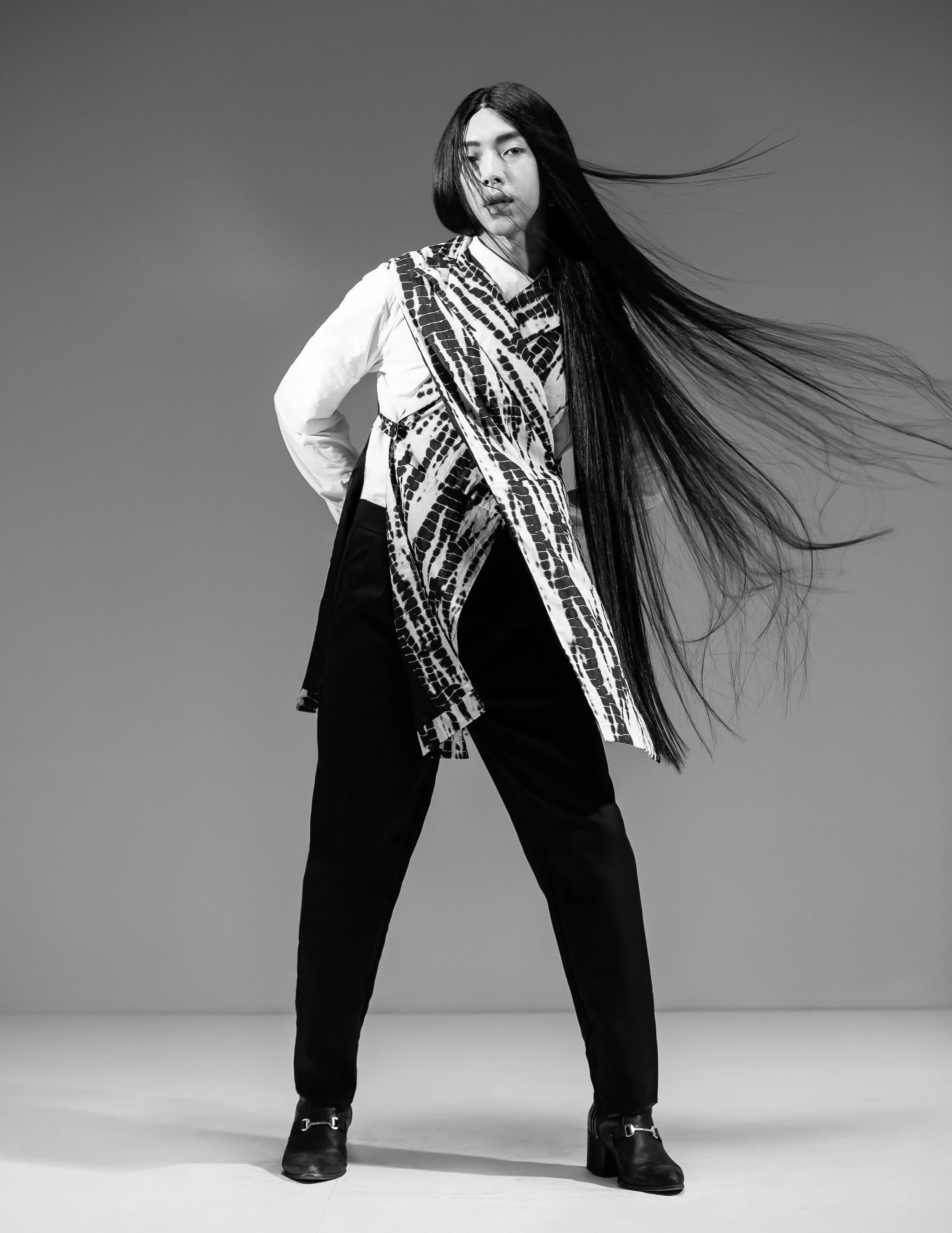 A black-and-white photograph of a person with long hair, standing with legs apart, against a plain background. They are wearing dark pants, boots, a patterned scarf, and a white shirt. They have a confident stance and their hair is flowing to the rig