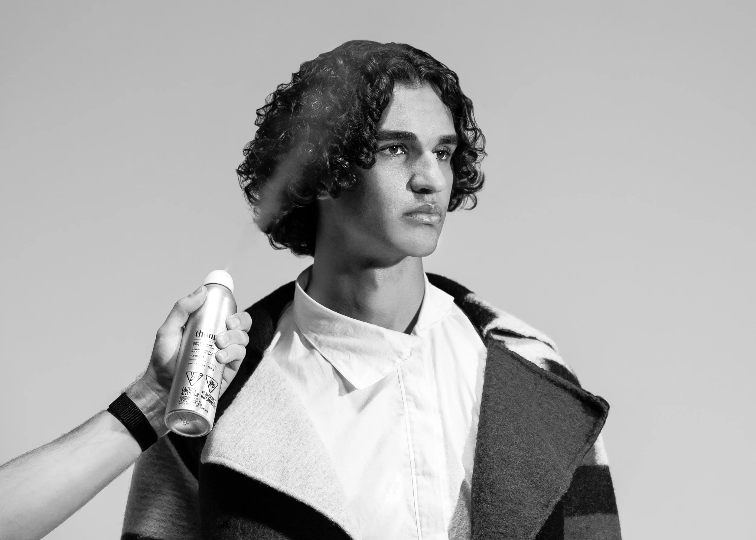 Black and white photo of a young man with curly hair wearing a white shirt and a dark coat draped over his shoulders, holding a can of spray in one hand.