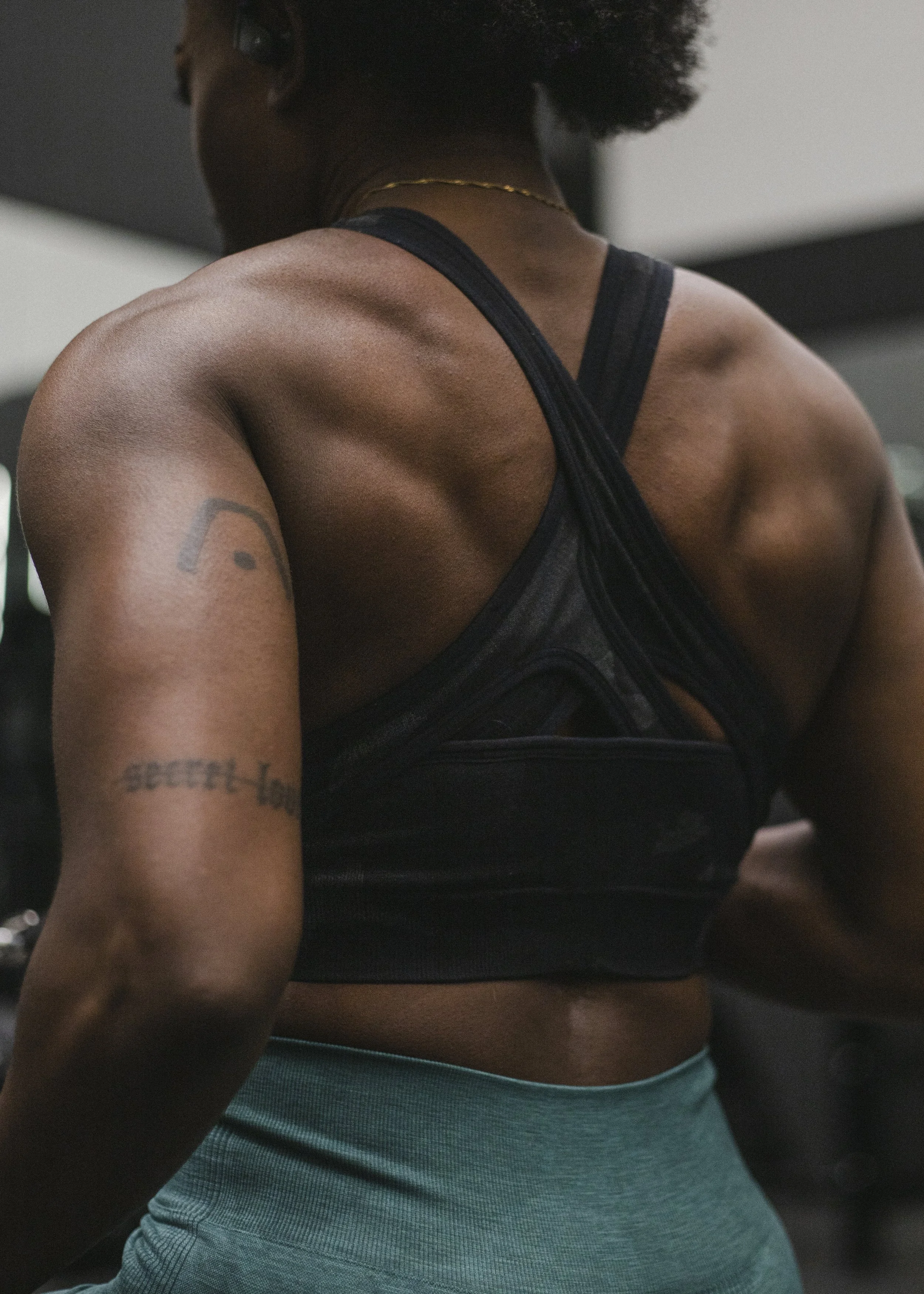 Close-up of an athletic woman with dark skin, wearing a black sports top and teal workout pants, with tattoos on her arm, in a gym environment.