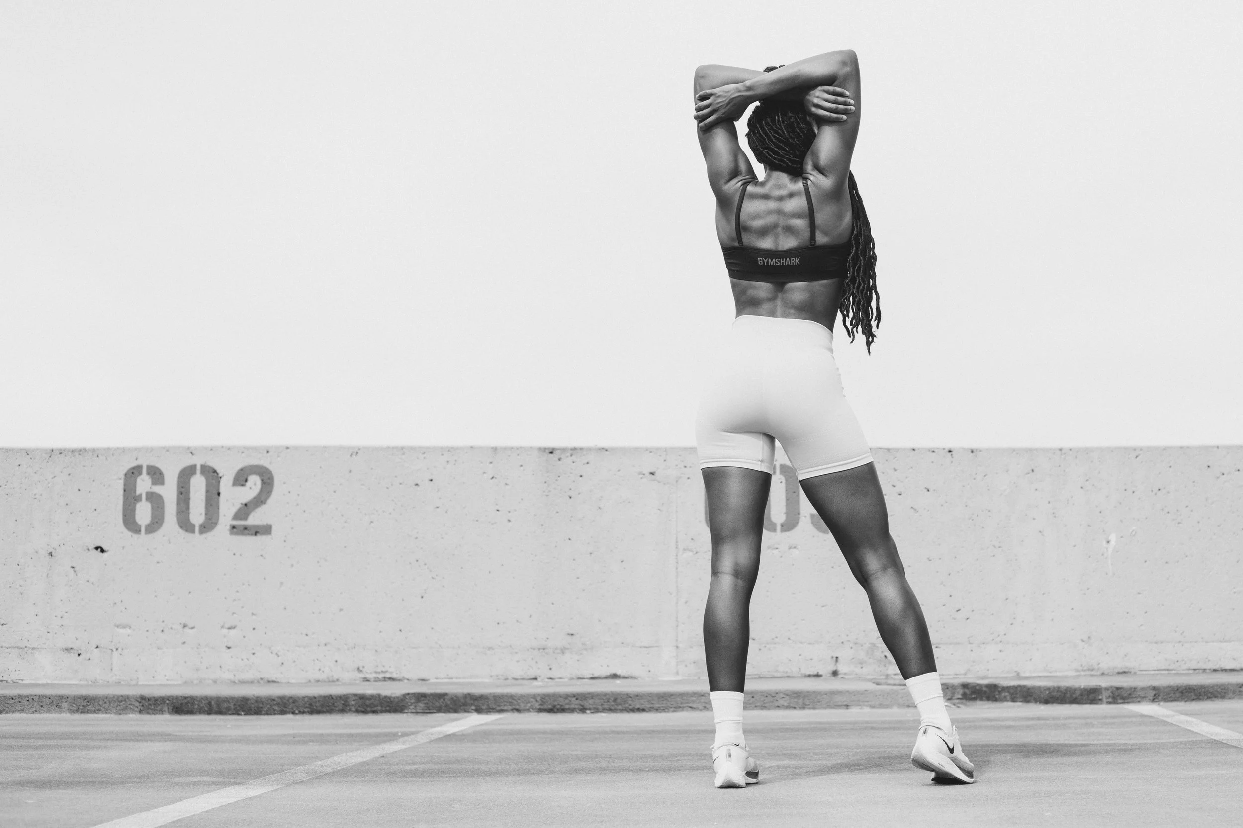 A woman with long dreadlocks stretching, wearing a sports bra and shorts, standing on a parking lot in front of a concrete wall with a height measurement of 602. Possibly preparing for or ending a workout.