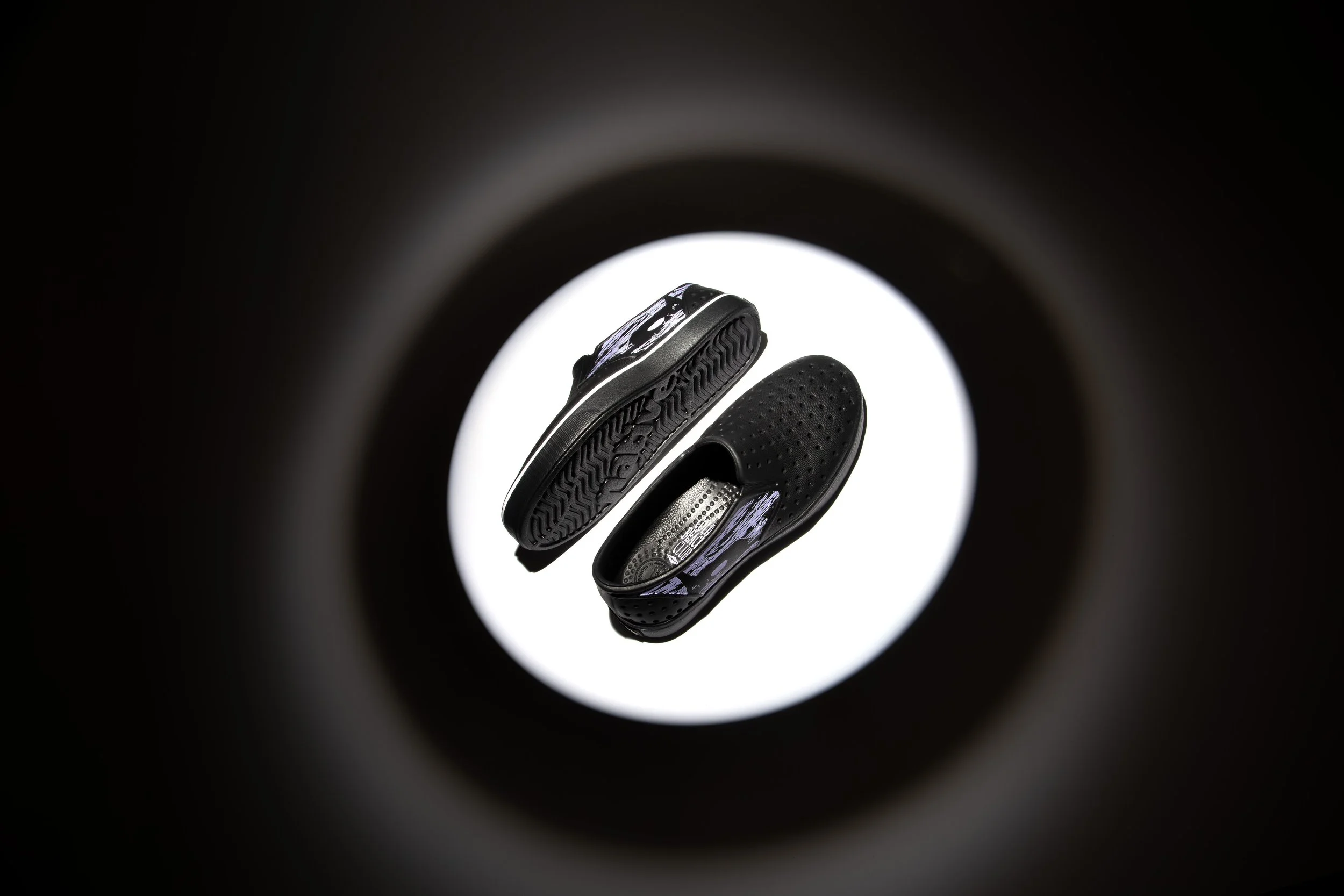 A black slip-on sandal with perforations, shown from above in a circular frame with a white background.
