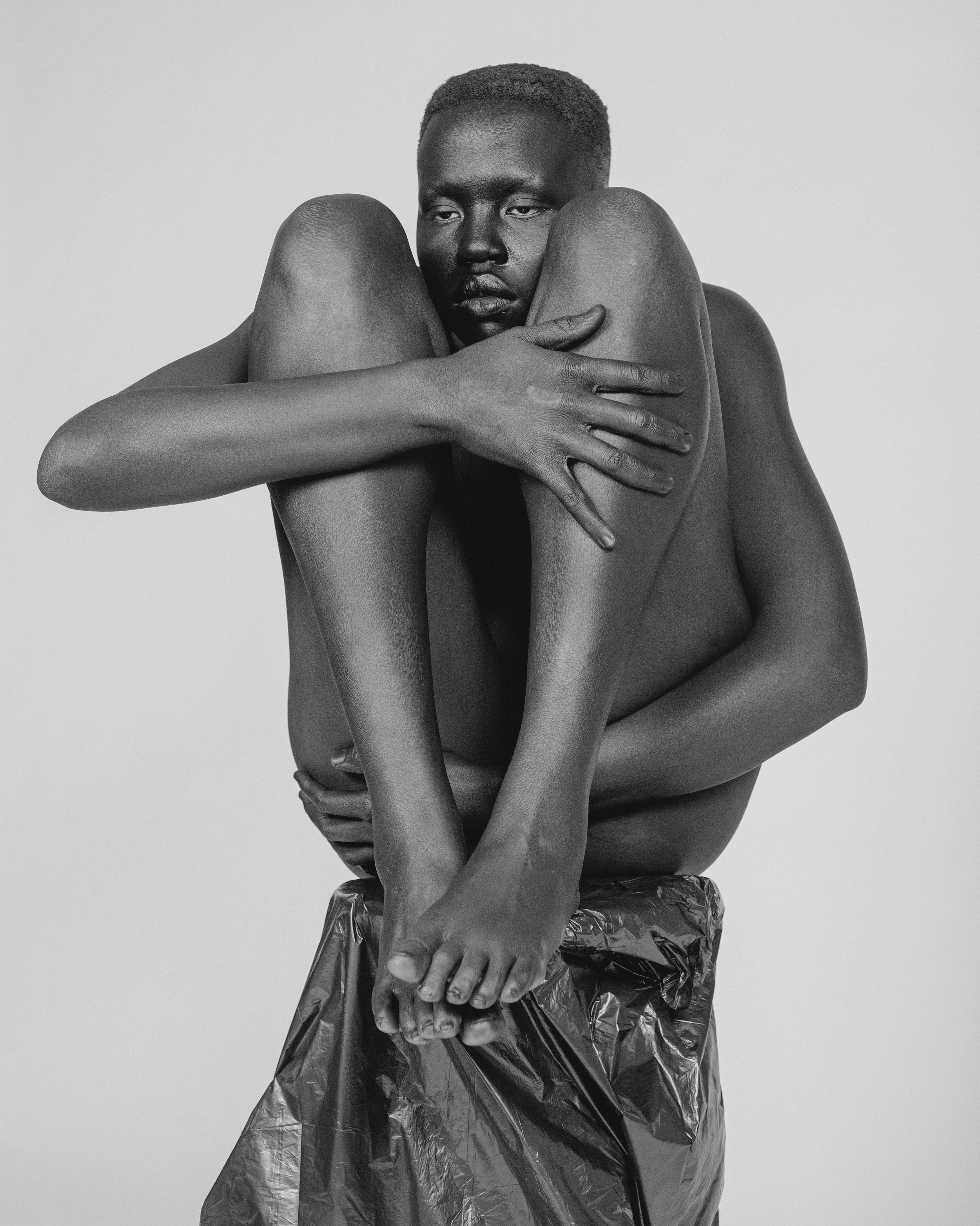 Black and white photo of a person with short hair, sitting cross-legged and hugging their knees on a plastic wrap against a plain background.