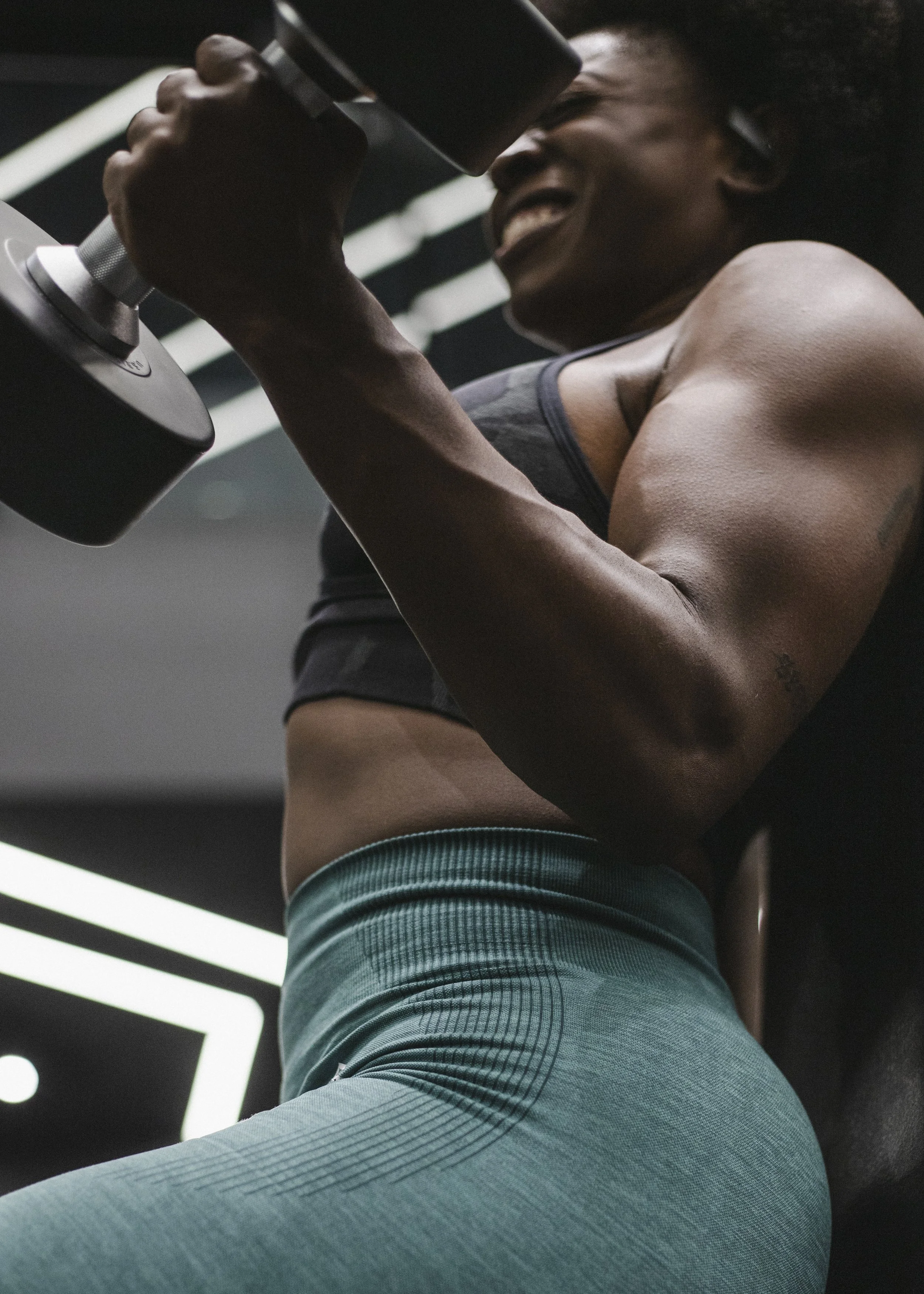 A person with dark skin and curly hair, wearing a gray athletic top and teal pants, is lifting a dumbbell at the gym, smiling with their eyes closed.