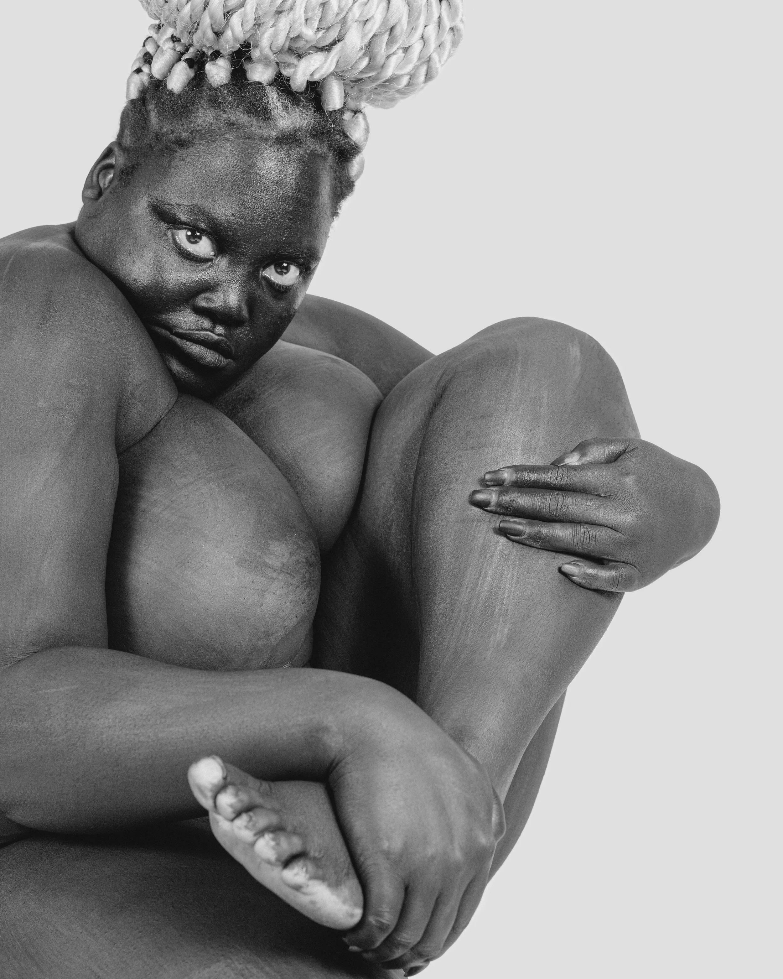 Black and white photo of a person with light-colored dreadlocks, looking at the camera, hugging their knees close to their chest.