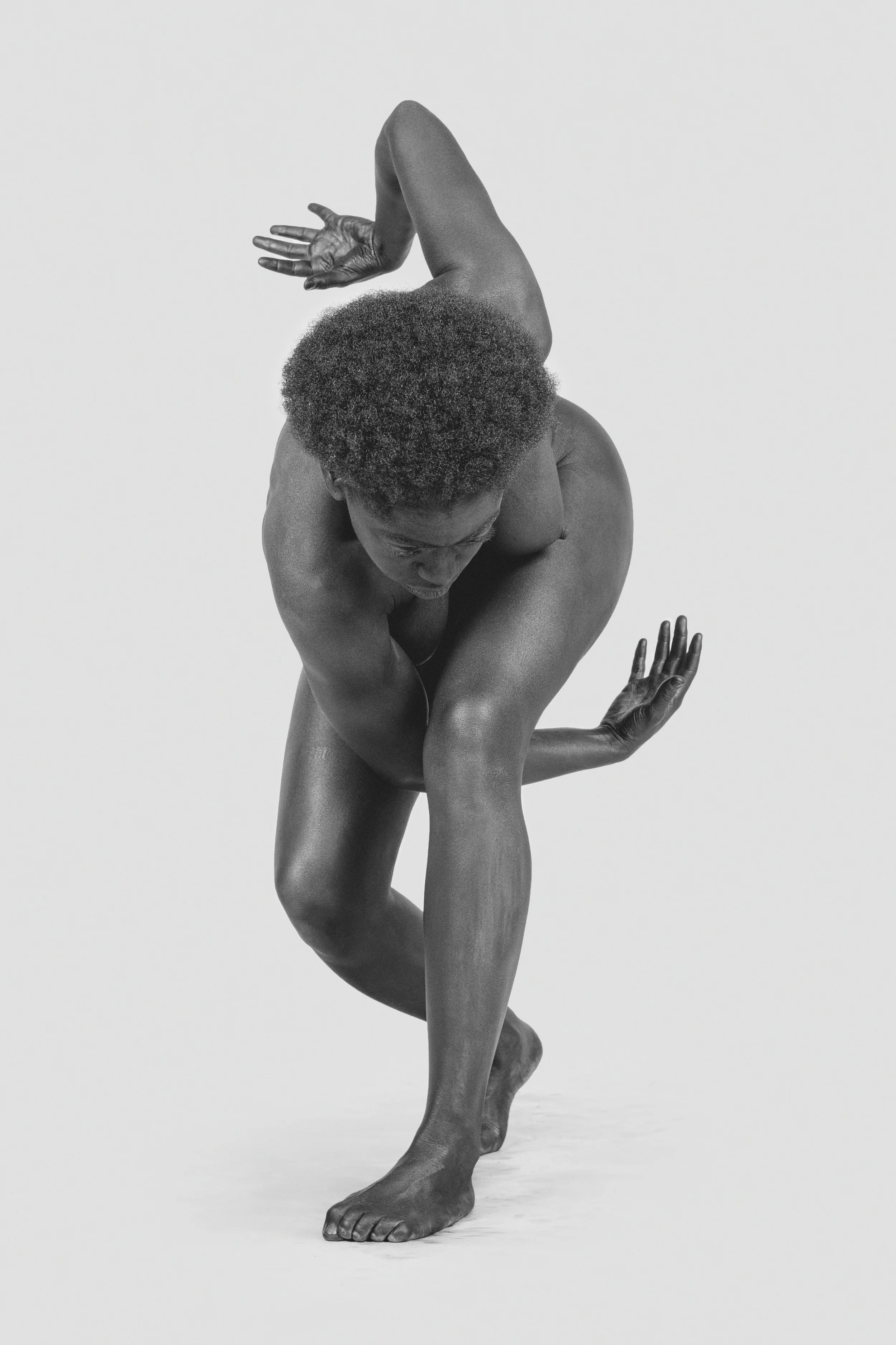 Black and white photo of a person with dark skin balancing on one foot, with their hair in a natural afro, arms raised, and hands gracefully positioned, against a plain background.