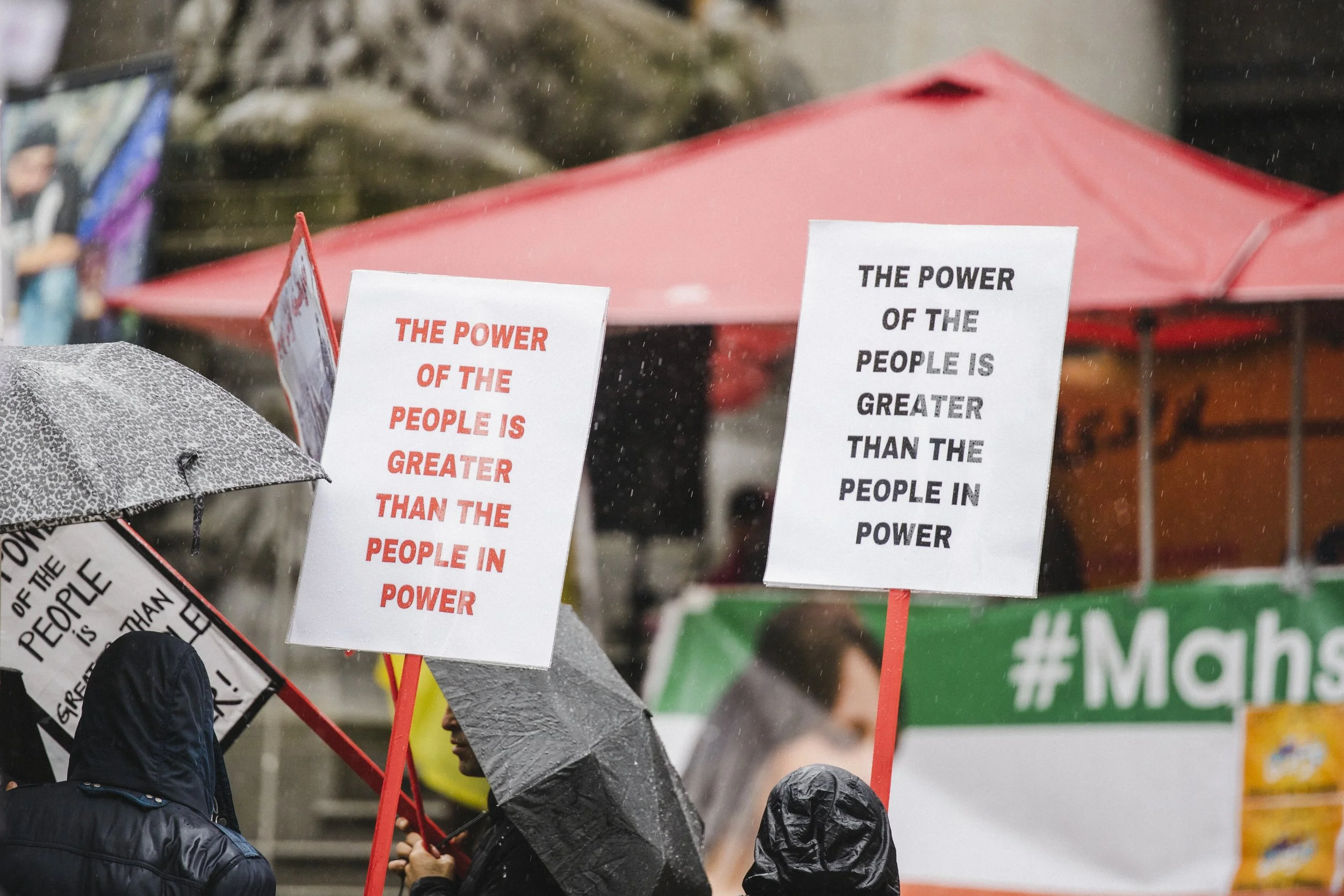 Protesters holding signs that read 'The power of the people is greater than the people in power' in a rainy outdoor setting, with umbrellas and a red canopy in the background.