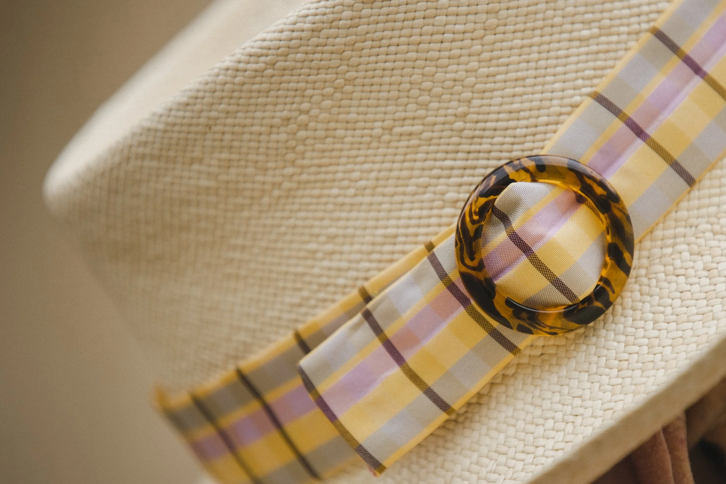 Close-up of a yellow, pink, and brown plaid fabric with a tortoise shell button on top of a cream woven straw hat.