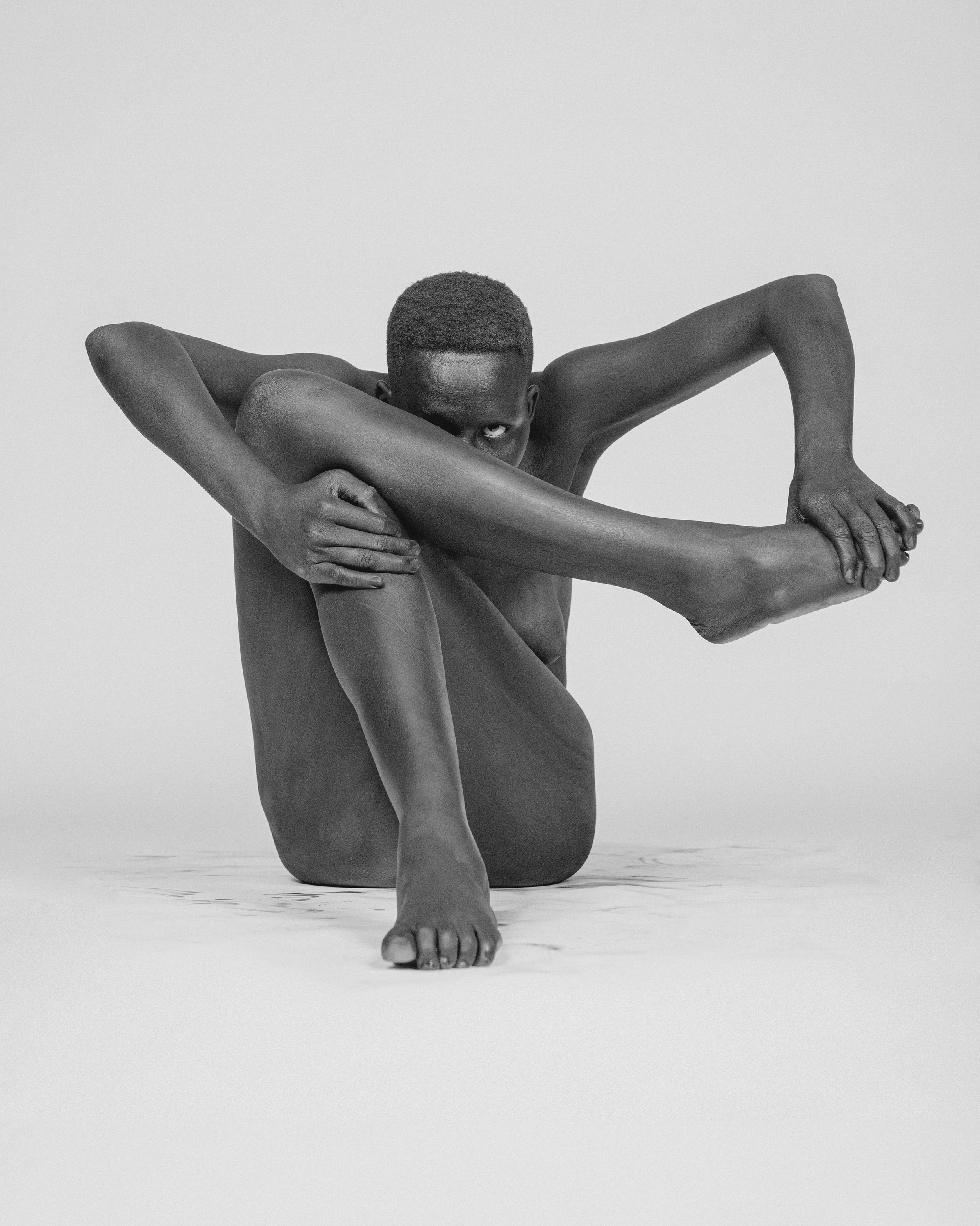 Black and white photograph of a man with a short haircut and minimal clothing, kneeling on the floor with his arms wrapped around his legs and looking directly at the camera.