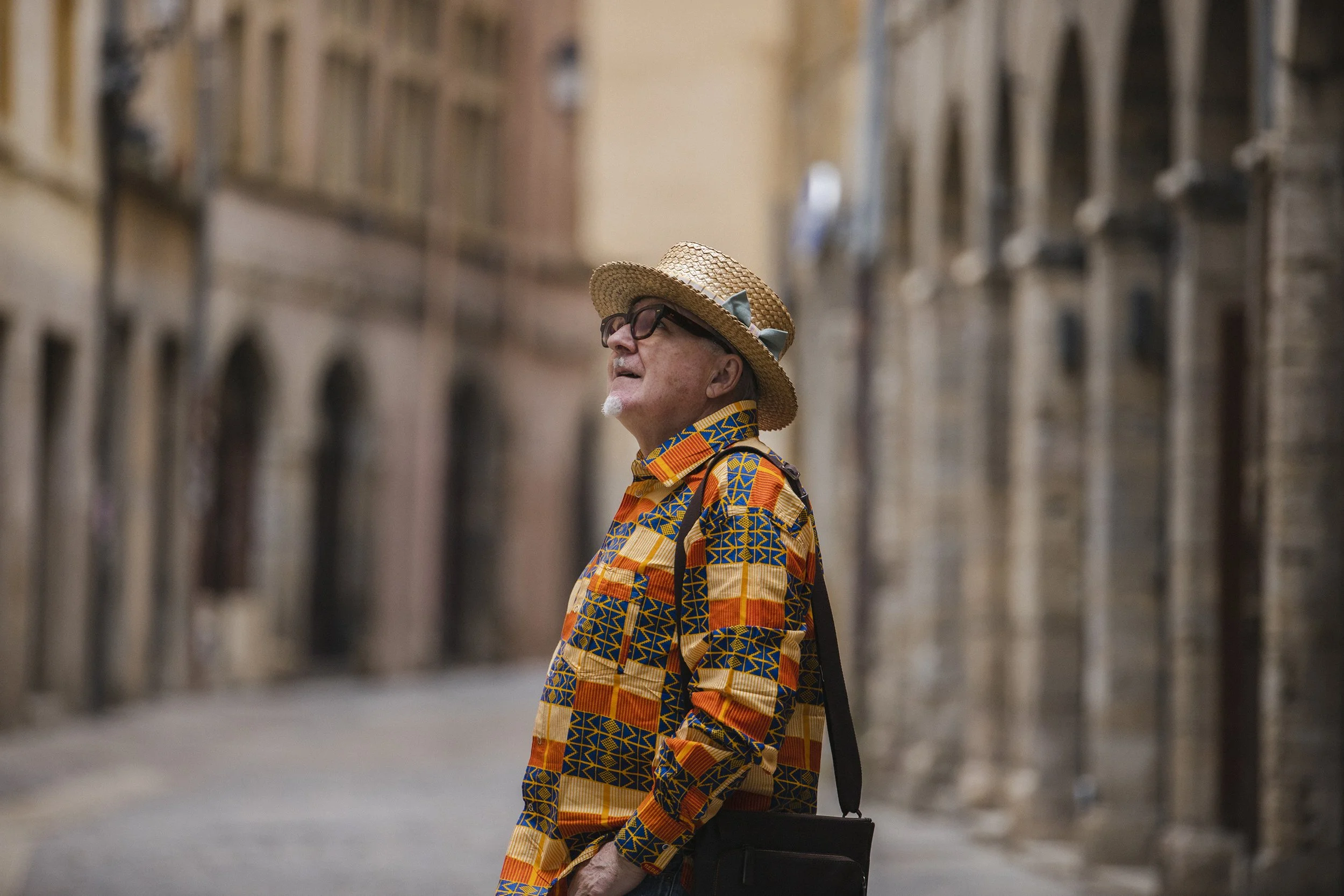 An elderly man wearing a straw hat, glasses, and a colorful, patterned shirt stands in a narrow street lined with buildings.