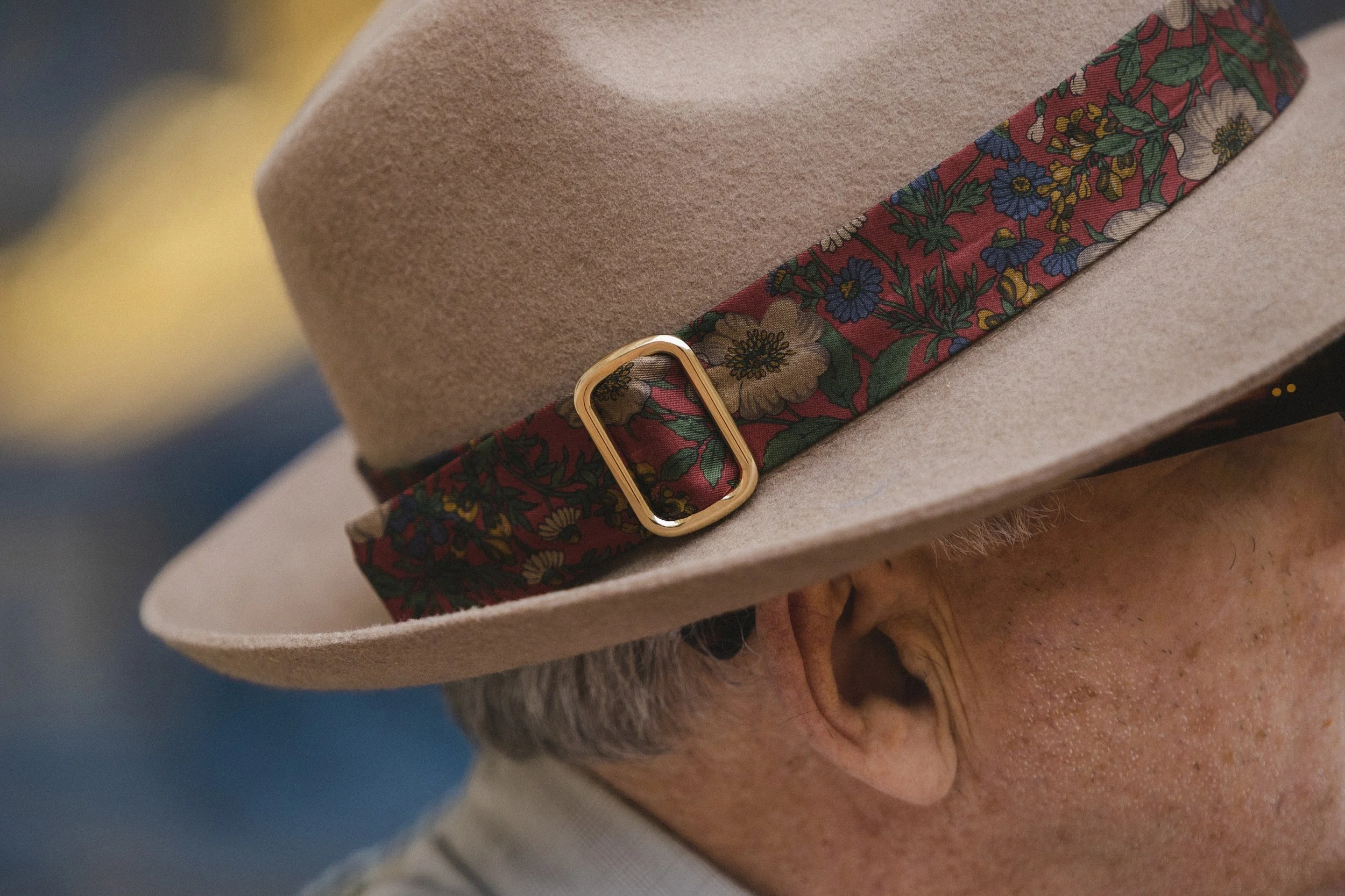 Close-up of a person's head wearing a beige felt hat with a decorative band featuring a floral pattern and a gold buckle.