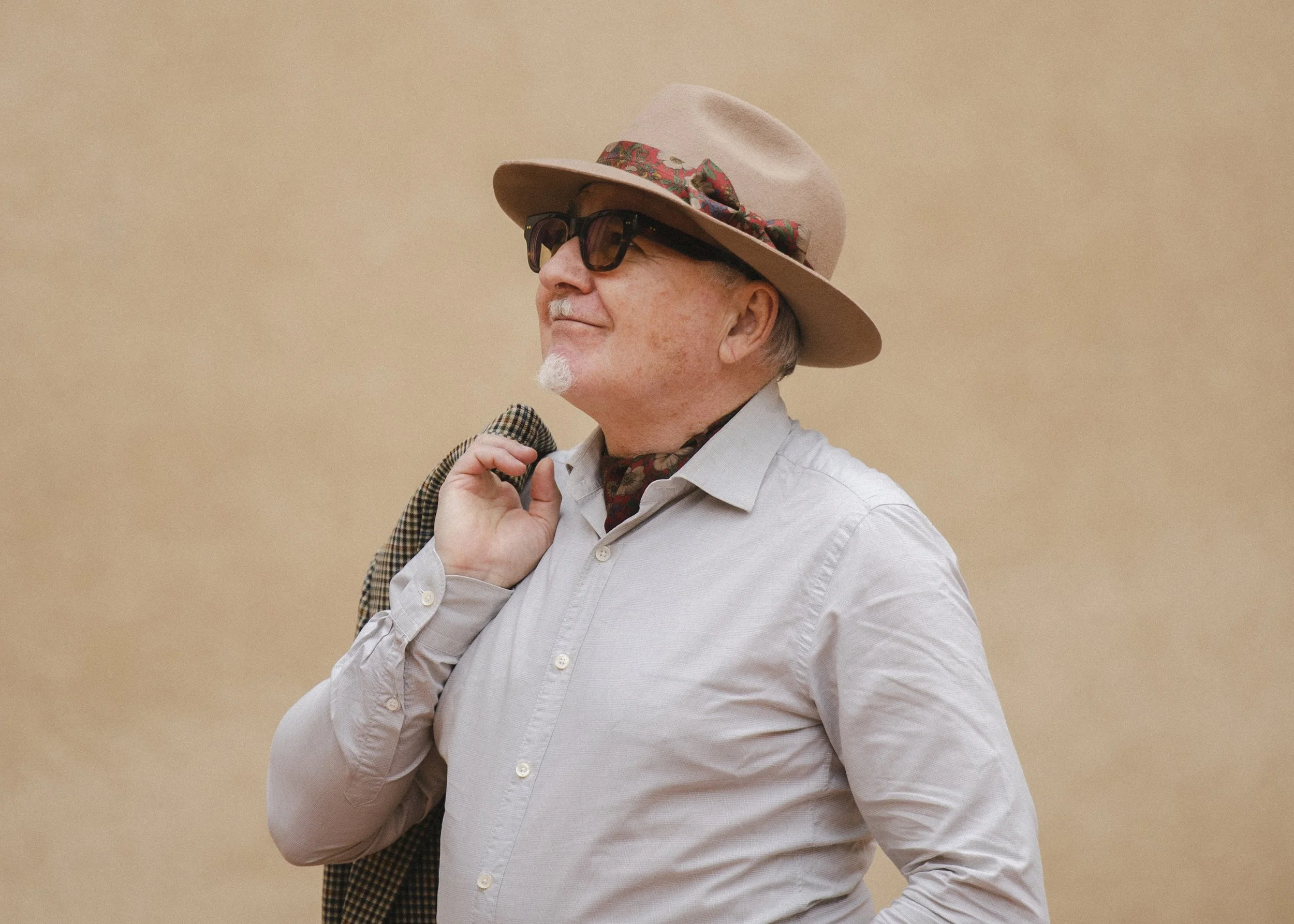 A man with light skin, gray hair, and a white beard wearing dark sunglasses, a light gray button-up shirt, a beige fedora hat with a floral ribbon, and holding a plaid jacket over his shoulder against a beige background.