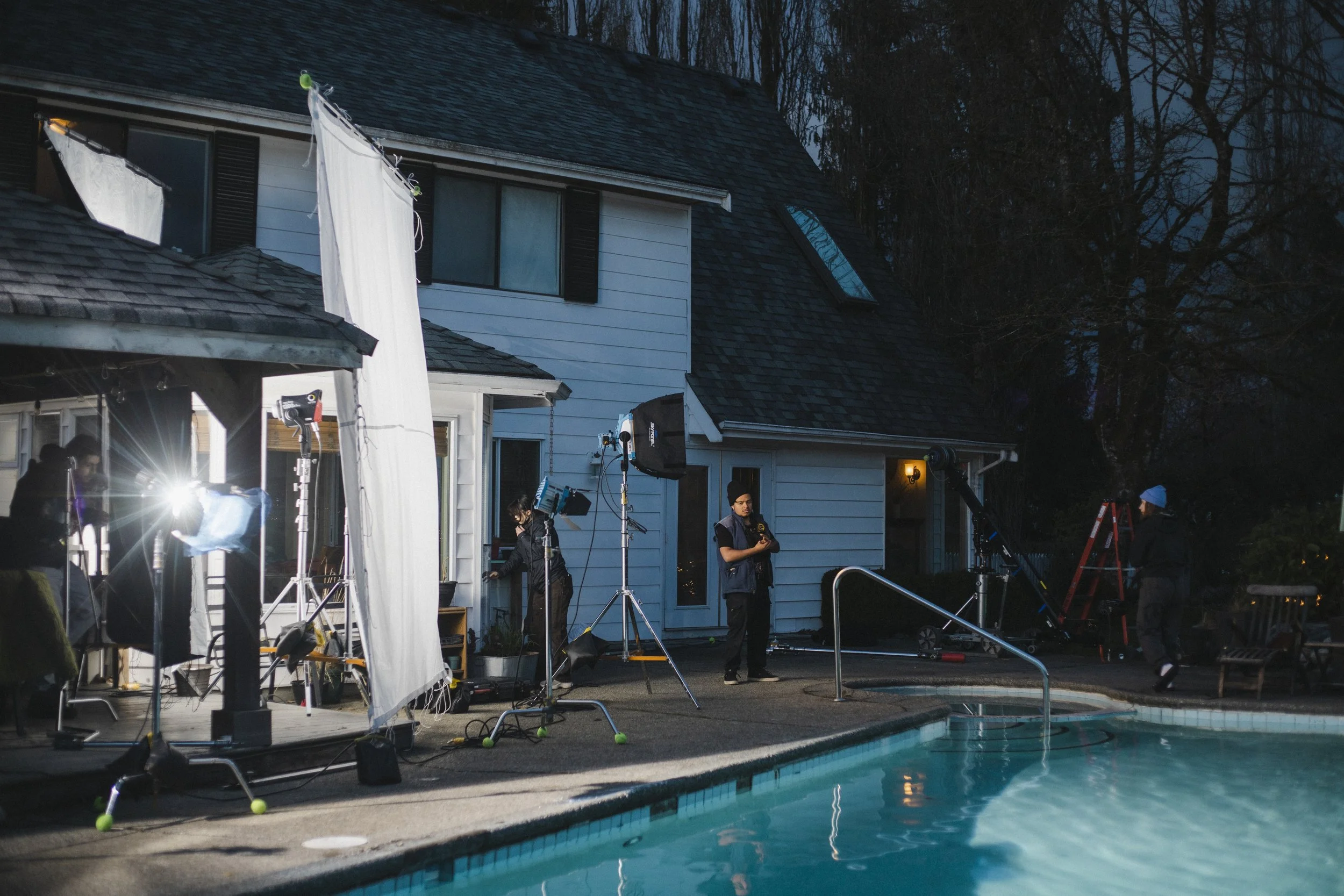 Film crew setting up lights and equipment outdoors near a swimming pool and a suburban house at dusk.
