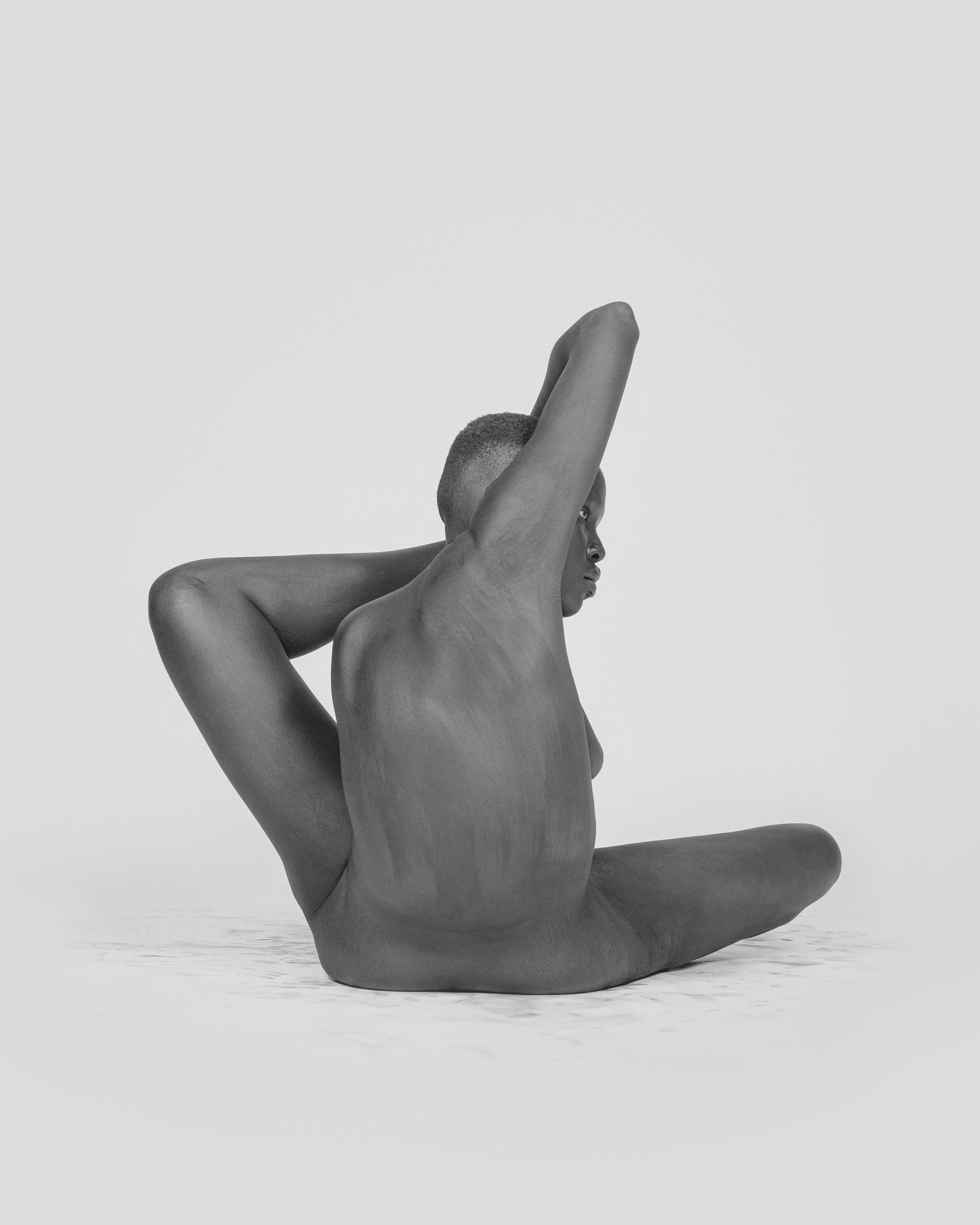 Black and white photograph of a person in a yoga pose, kneeling on the ground with one leg extended forward and the other bent behind, with arms raised and hands resting behind their head, against a plain white background.