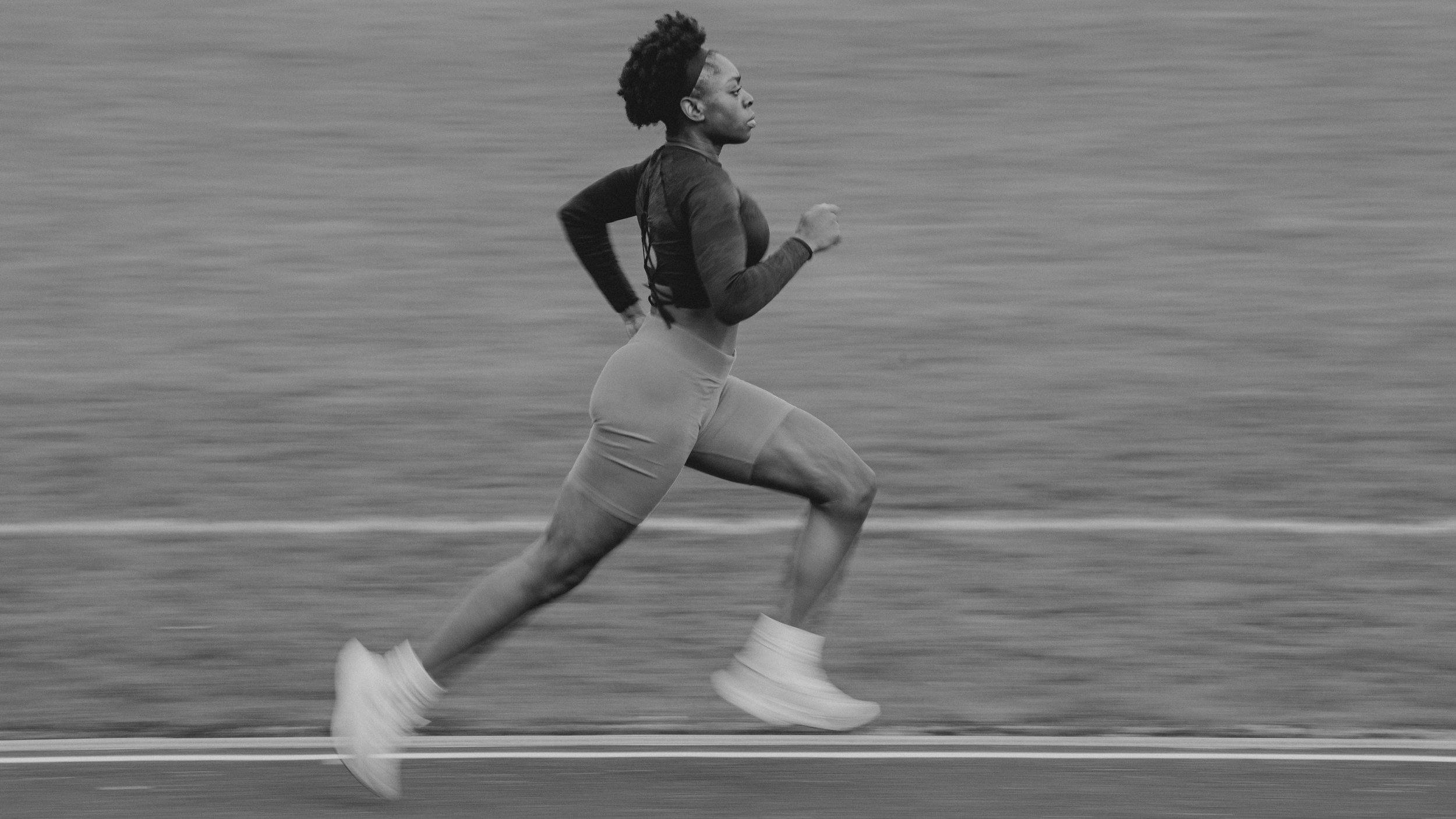 A woman running along a track near a body of water, dressed in athletic clothing and white sneakers, captured in black and white.