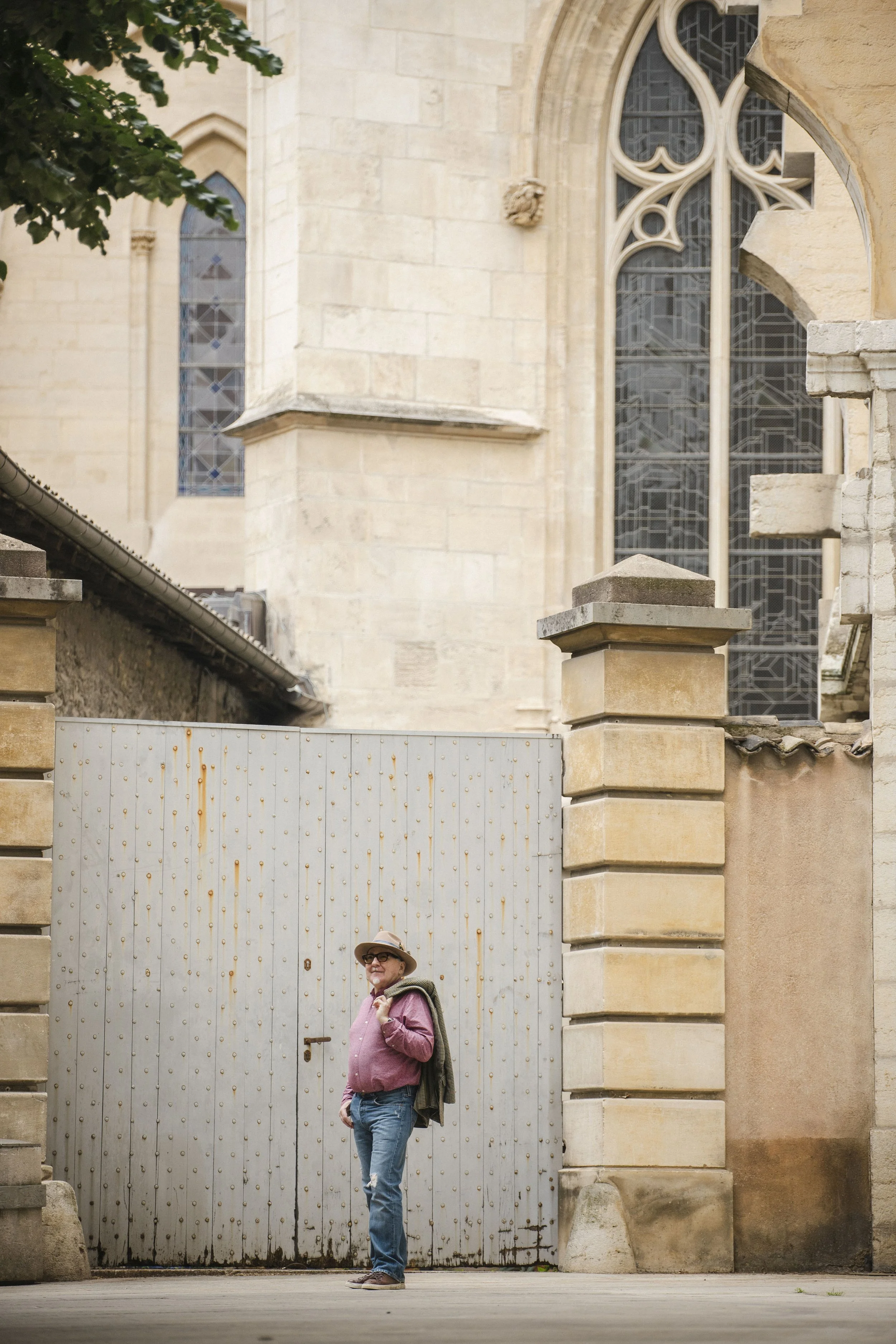 A woman wearing glasses, a wide-brimmed hat, a pink shirt, and jeans, standing in front of a large, wooden gate with metal accents, in front of an old stone church with stained glass windows, a stone wall, and a column.