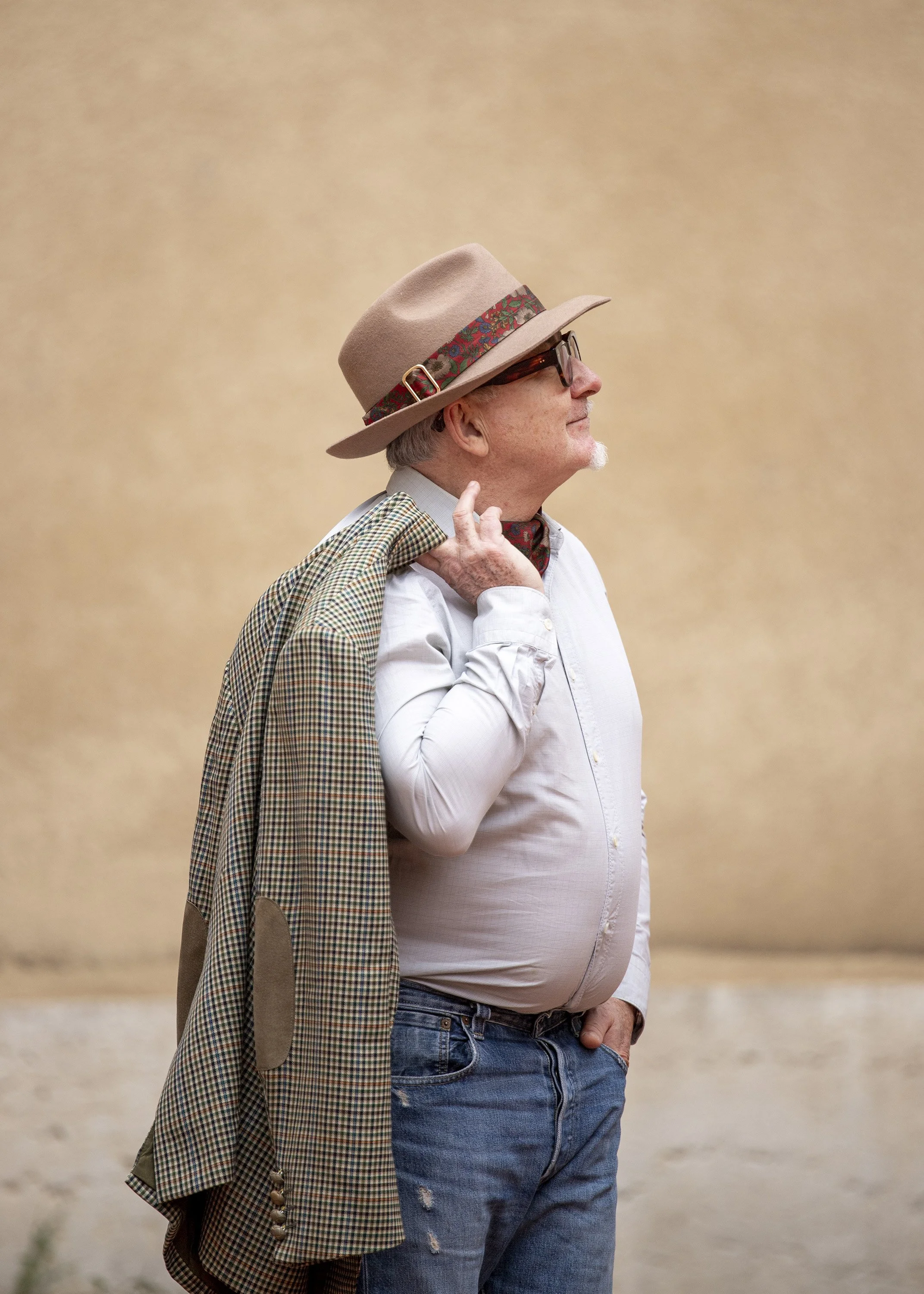 A man in a white shirt and jeans, wearing sunglasses and a felt hat with a floral band, stands outdoors against a beige background, holding a checked blazer over his shoulder with his right hand.