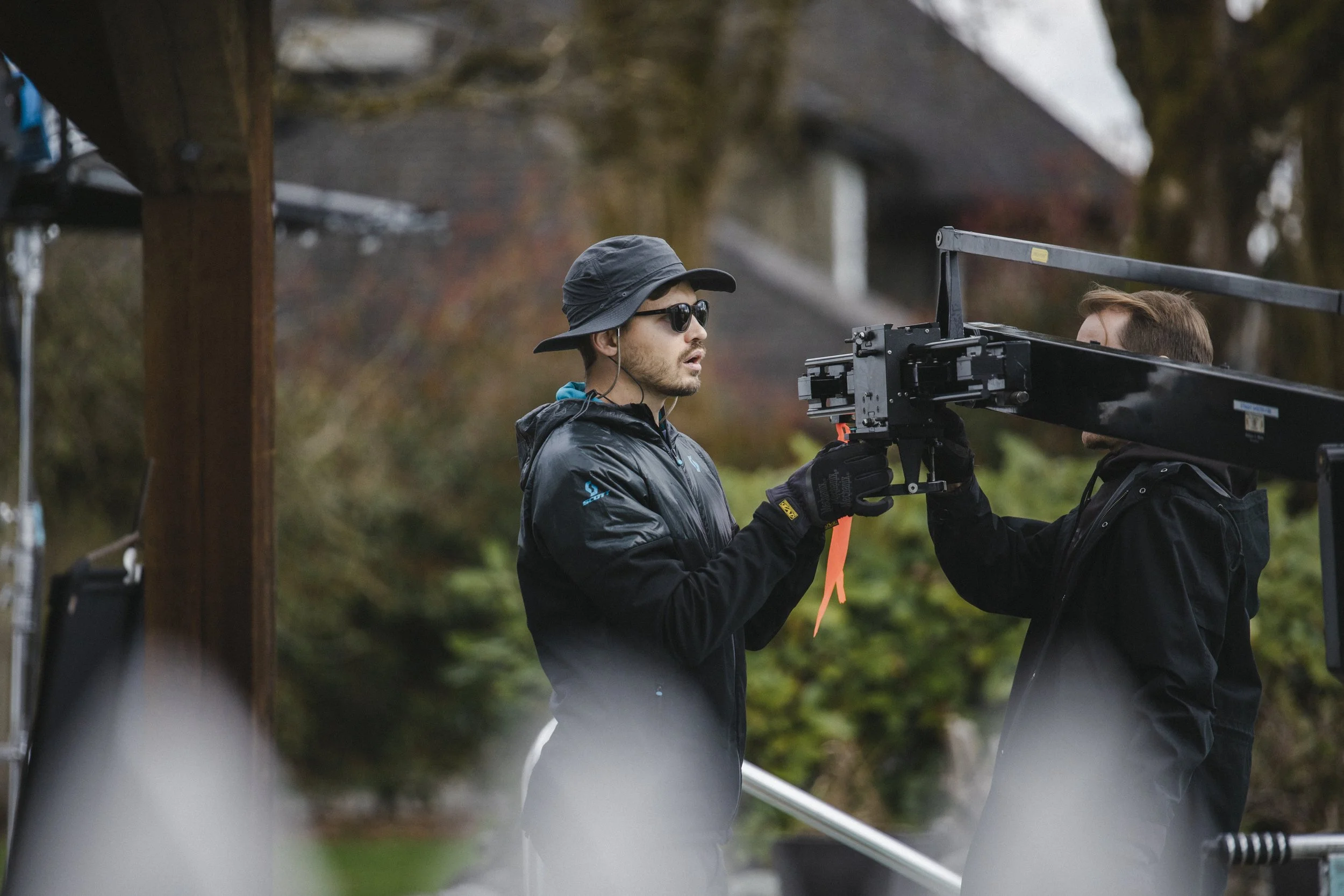 Two individuals on a film set, one wearing a black bucket hat, sunglasses, and a black jacket, and the other in a black hoodie, working with film equipment outdoors during daytime with a blurred background of trees and a building.