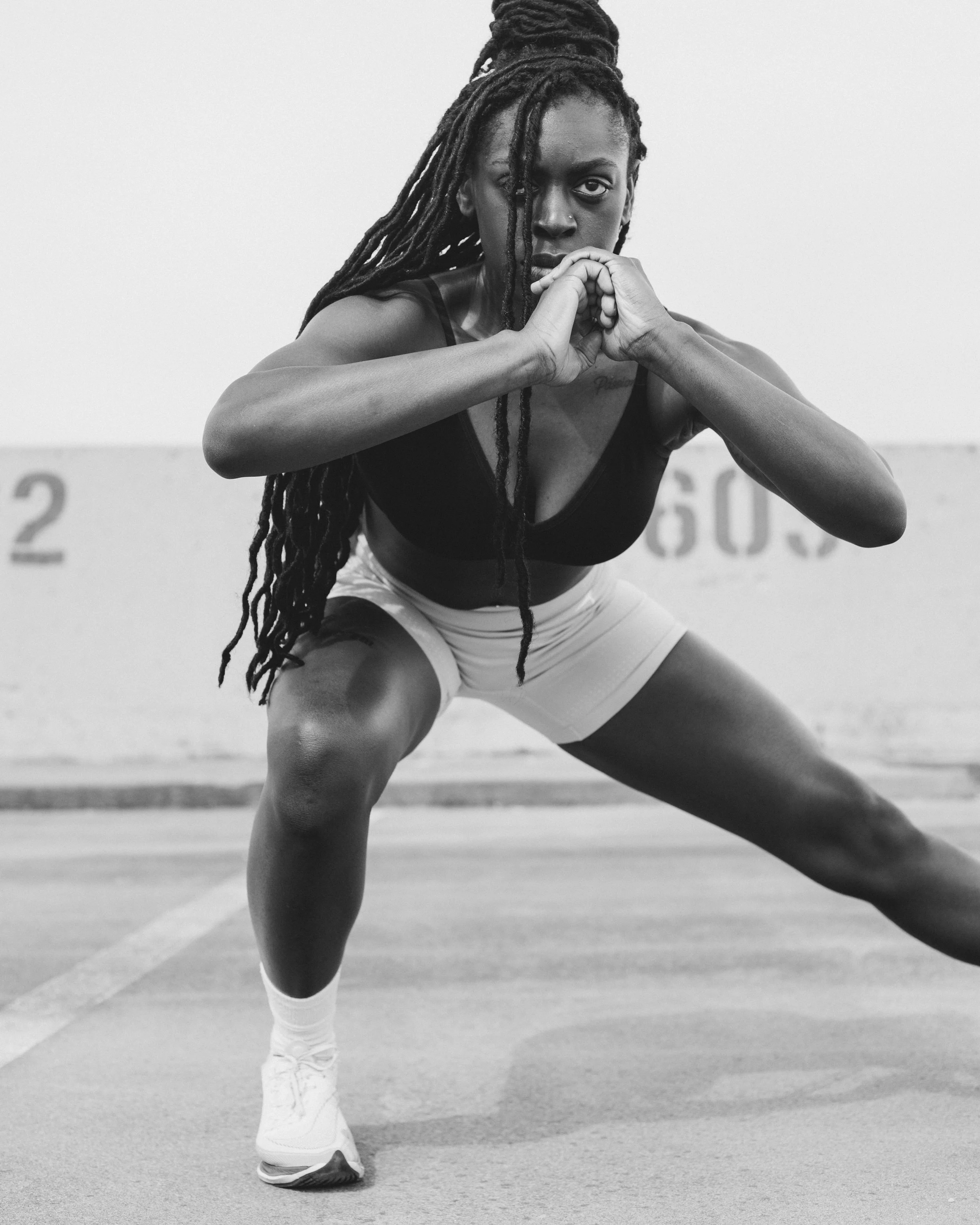 Black and white photo of a female athlete in a squat position, on an indoor track, with her hands clasped under her chin, wearing athletic gear and sneakers, facing the camera with an intense expression.