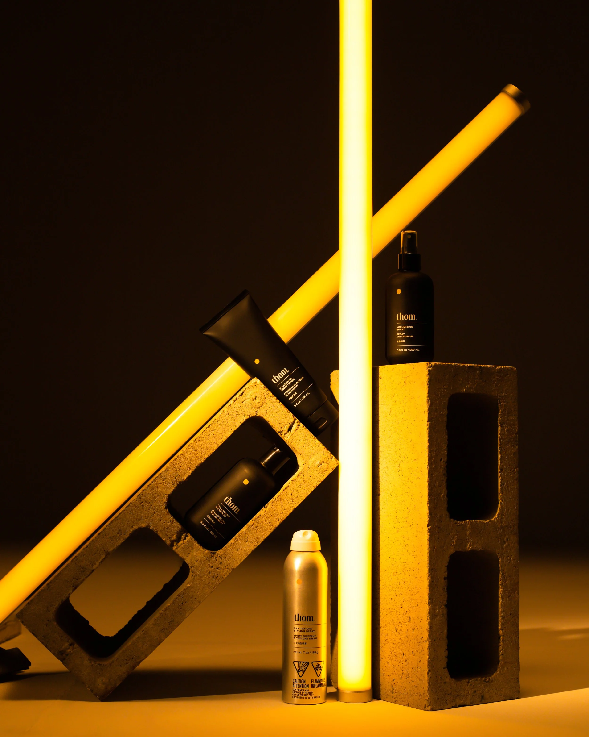 A black spray can labeled 'thom.' with caution and flammable warning symbols, a beige metallic spray bottle, illuminated yellow light tubes, a concrete block, and dark background.