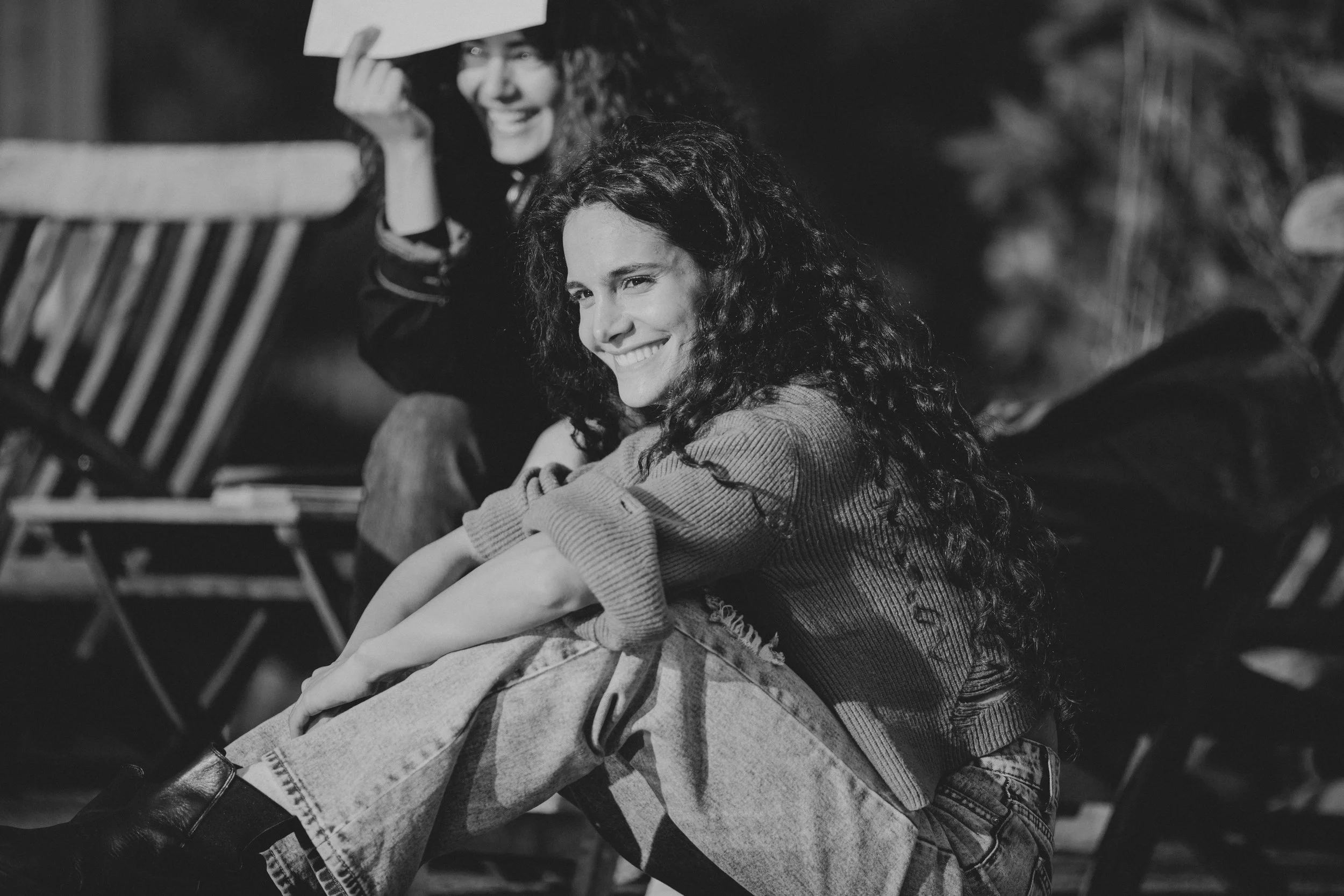 Black and white photo of two women smiling, sitting outdoors on wooden chairs, one wearing a hat and the other with long curly hair.