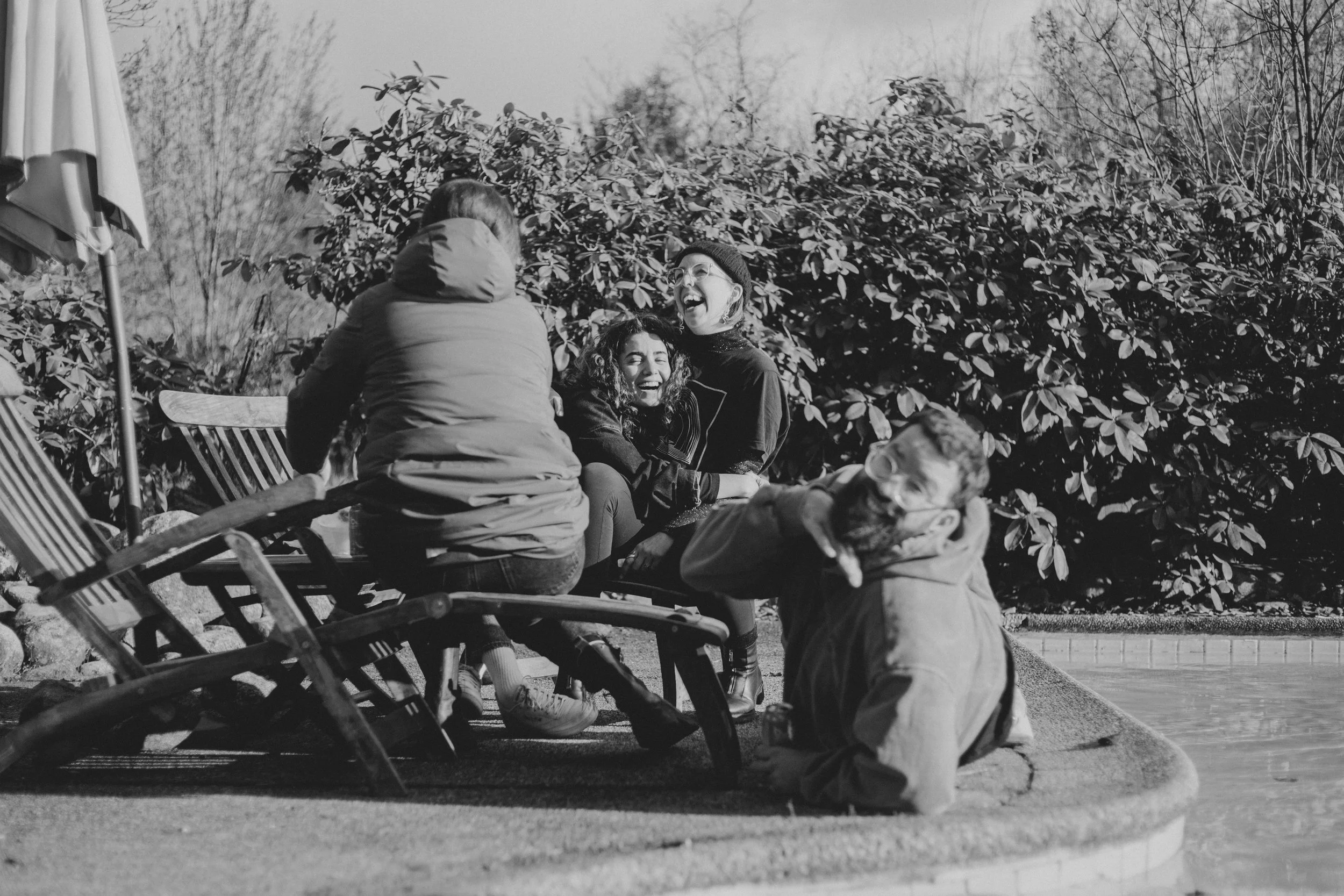 A group of people sitting outdoors in front of bushes, including a man, two women, and a child, enjoying a sunny day and laughing.