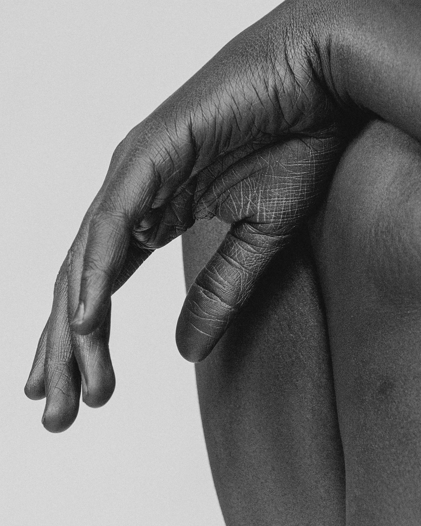 A black and white close-up photograph of a person's hand and shoulder, showing detailed skin texture and fine lines, with the hand gently touching the shoulder.