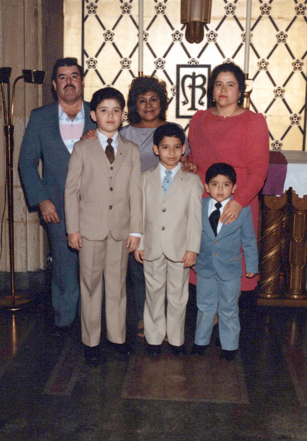 Family portrait of three adults and three children dressed in formal attire, standing indoors with decorative wall and religious symbols in the background.
