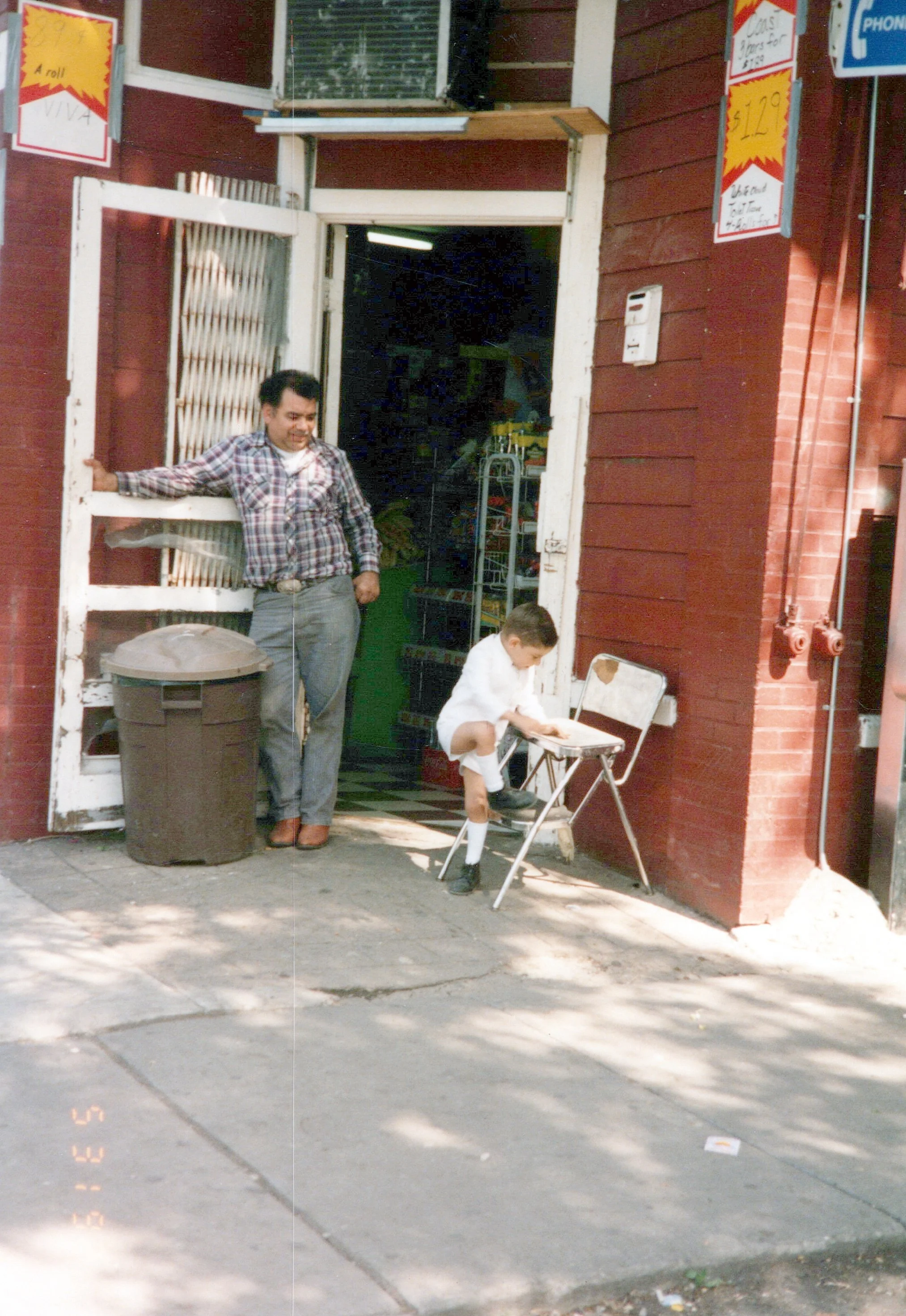 A man in a plaid shirt standing outside near a yellow trash can and a young boy in white sitting on a folding chair, with a red wooden building behind them featuring signs and a partially open door.