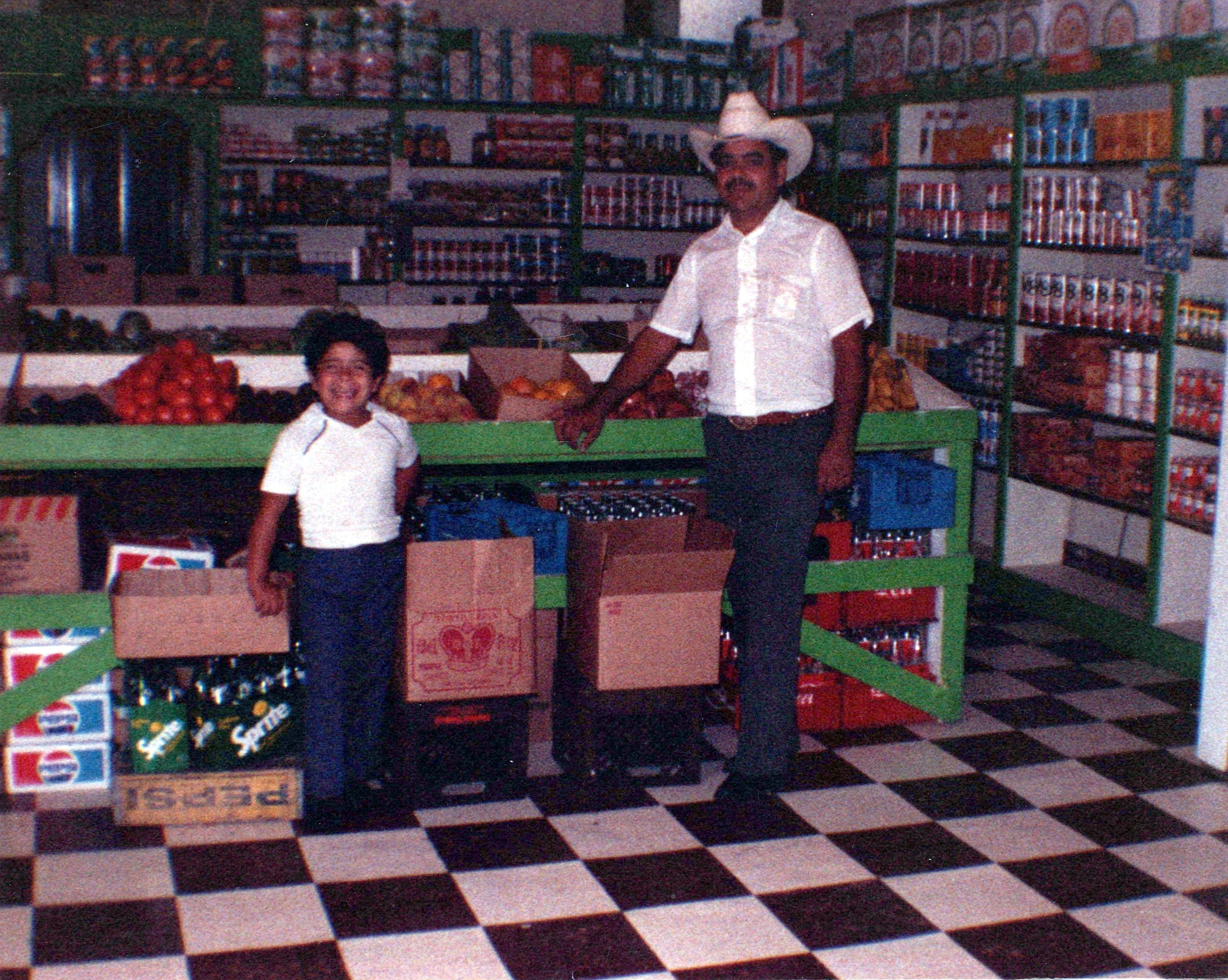 A man with a mustache, wearing a white cowboy hat and white shirt, is shopping in a grocery store. There is a young boy in a white shirt, standing in front of him. They are in front of shelves stocked with canned goods, boxes of chips, and other grocery items. The store has a black and white checkered floor.