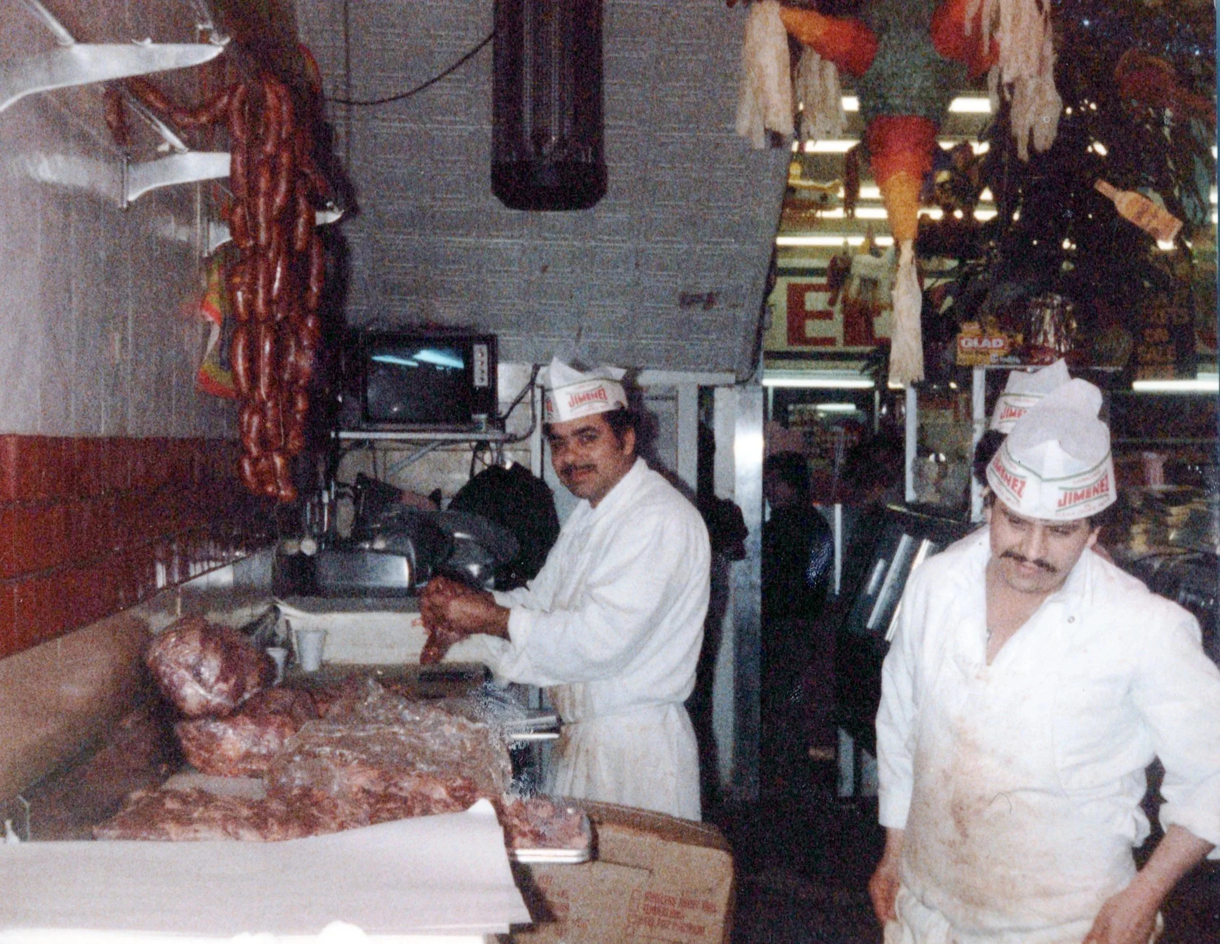 Kitchen scene with two men preparing meat, various meat cuts, onions, and kitchen utensils on a counter, and two men wearing aprons and hats working.
