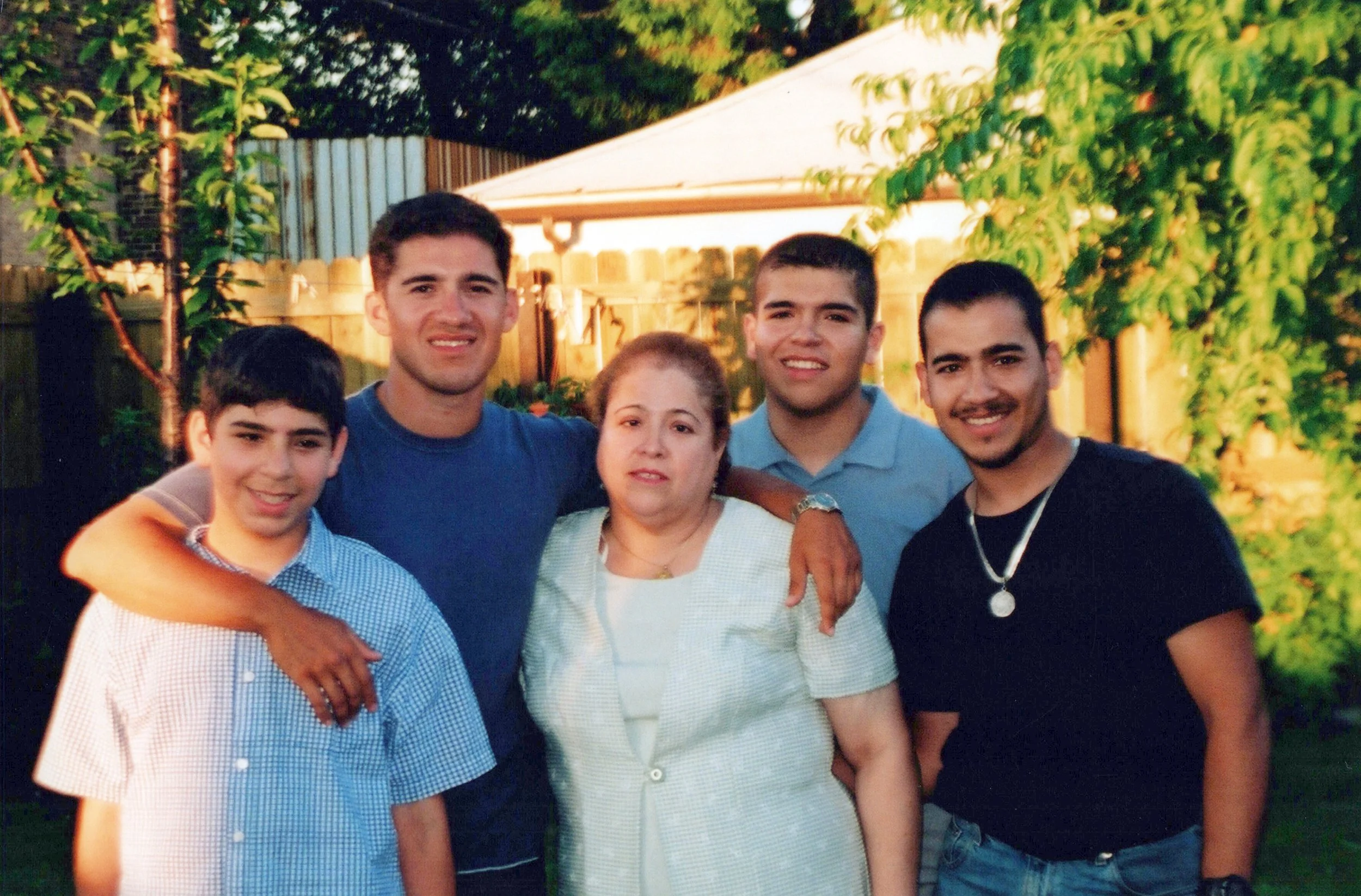 A group of six people, five young men and one older woman, standing close together outdoors during the daytime with a wooden fence, trees, and a house in the background. They are smiling and appear to be enjoying a gathering.