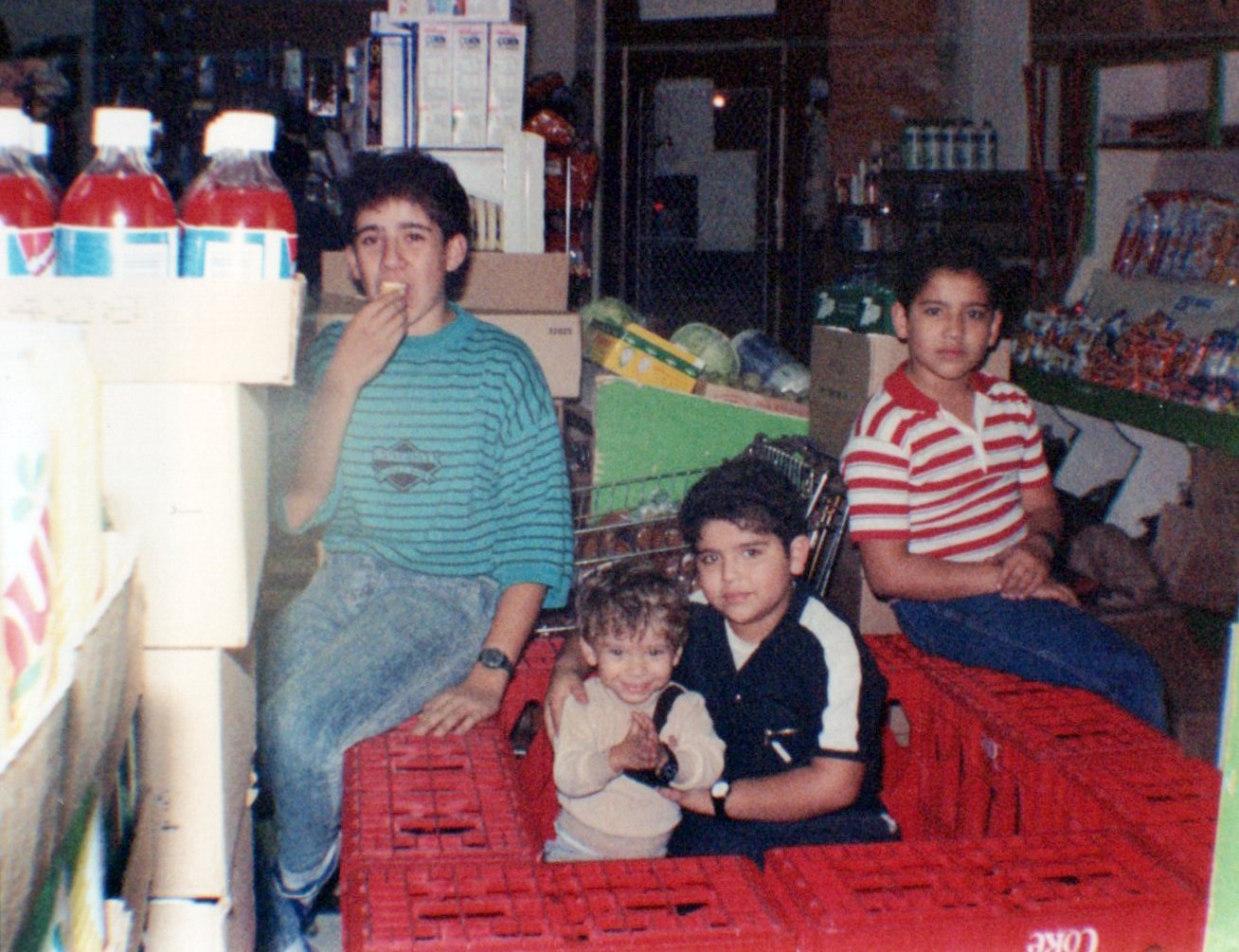 Four children inside a store, sitting on red crates, surrounded by shelves of snacks and groceries. One boy in a striped shirt is eating, and the others are posing for the camera.