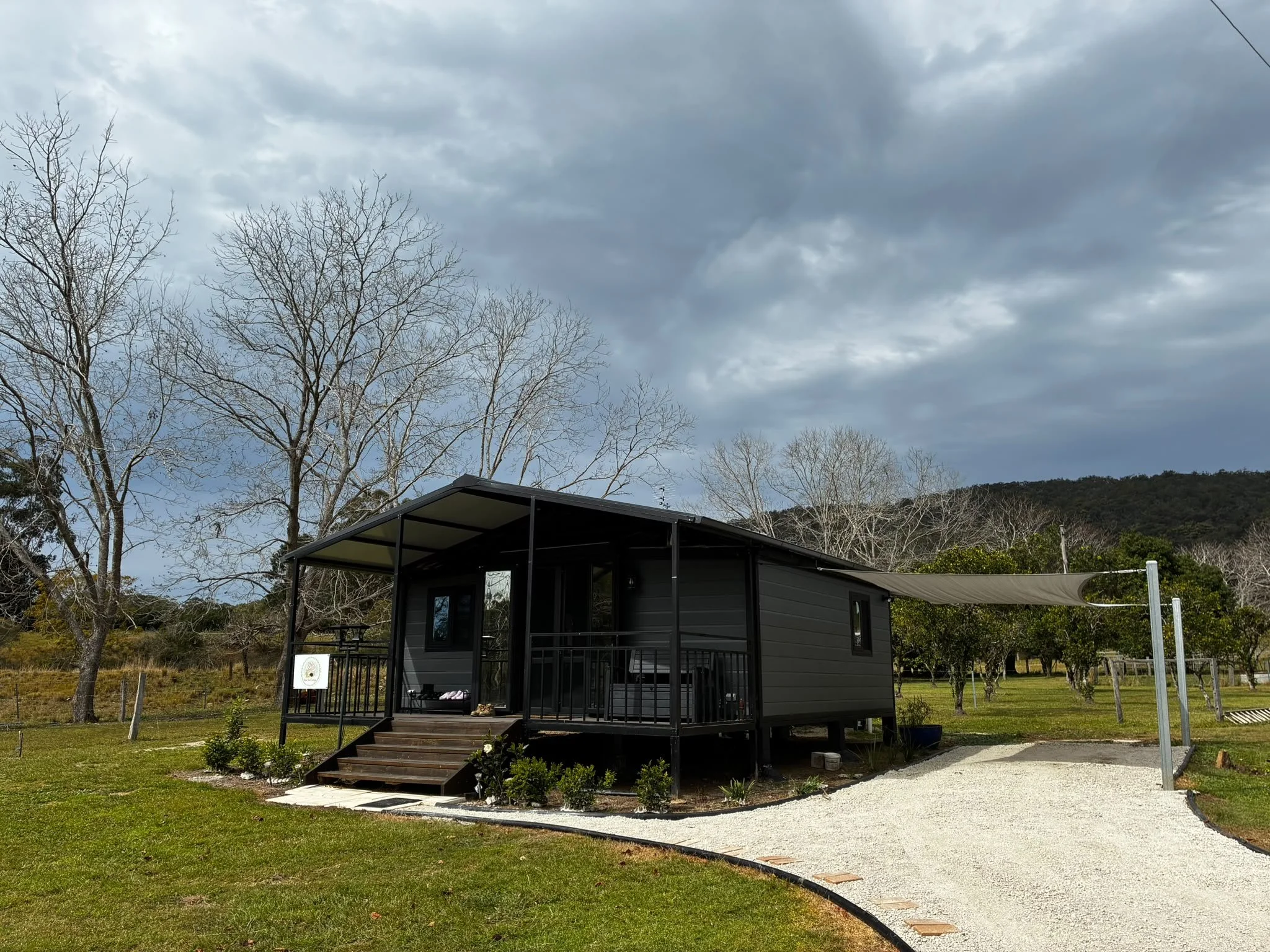 A small black house with a deck and stairs, surrounded by green grass and trees with bare branches, under a cloudy sky.