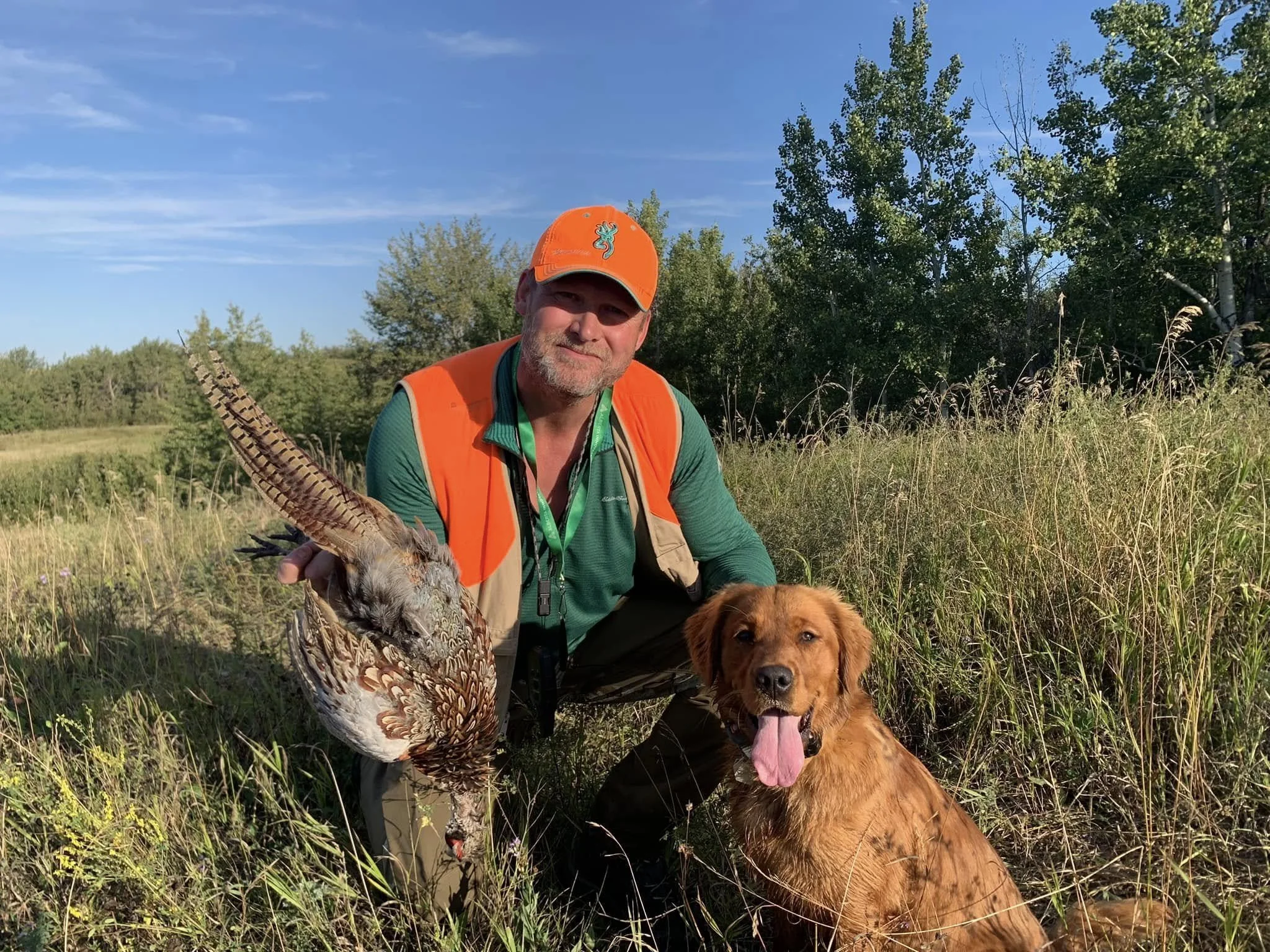 A man wearing an orange hunting cap and vest, holding a dead bird, with a golden retriever sitting beside him outdoors in tall grass and trees, under a blue sky.