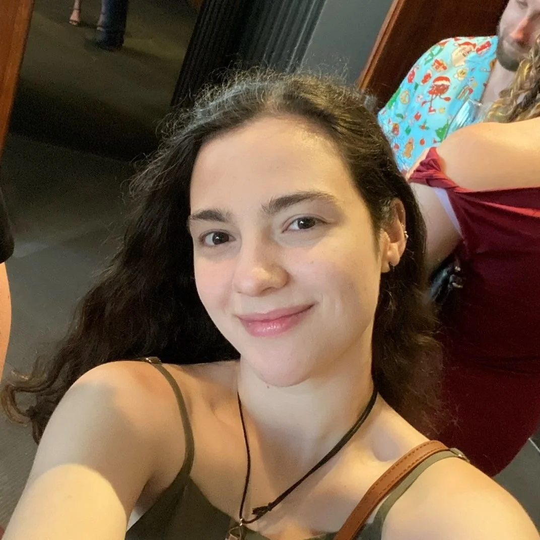 Young woman with dark, curly hair taking a selfie. She is smiling and wearing a black sleeveless top. In the background, there are glimpses of other people, including someone in a colorful shirt with a pattern and another in a red dress. Part of the indoor setting is visible.