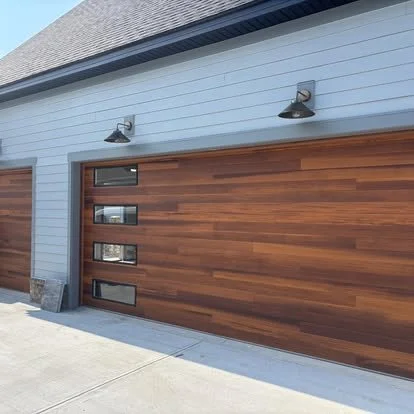 A modern garage door with a wood finish and three small horizontal windows on the left side, attached to a light blue house with exterior lights above the door.
