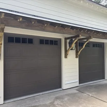 Two dark-colored garage doors with white siding and a small section of concrete driveway, with wooden braces supporting the roof overhang.