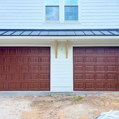 Close-up of a house's garage with two brown garage doors and a white exterior wall, featuring a window above, under a metal roof.